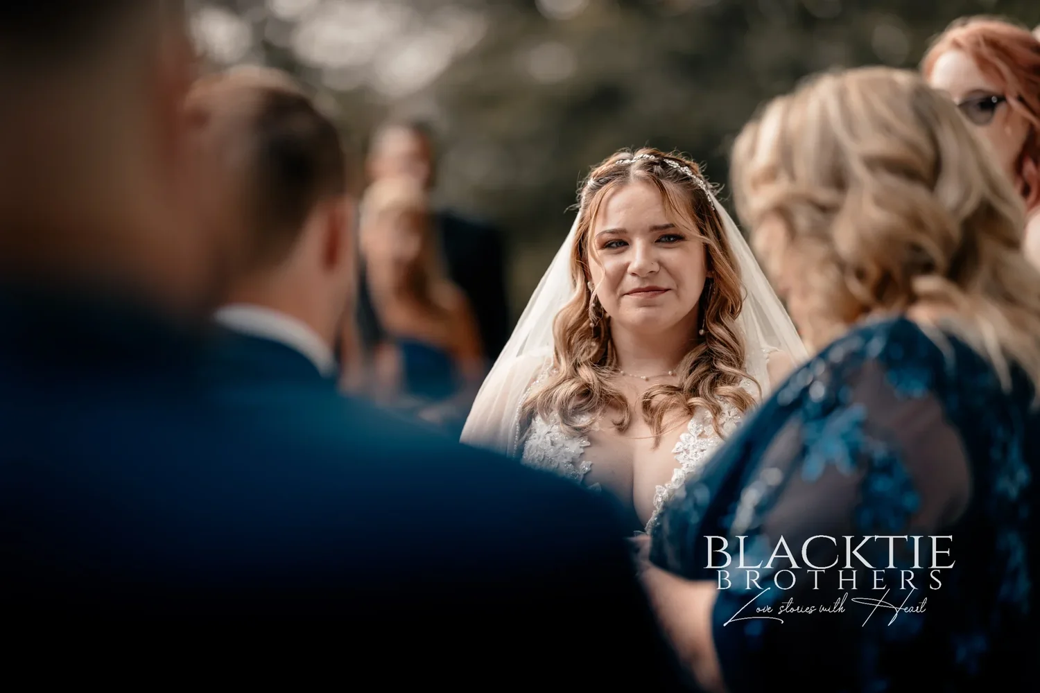 Jennifer under her veil looking at Jared during the ceremony, the officiant and a bridesmaid softly out of focus around her