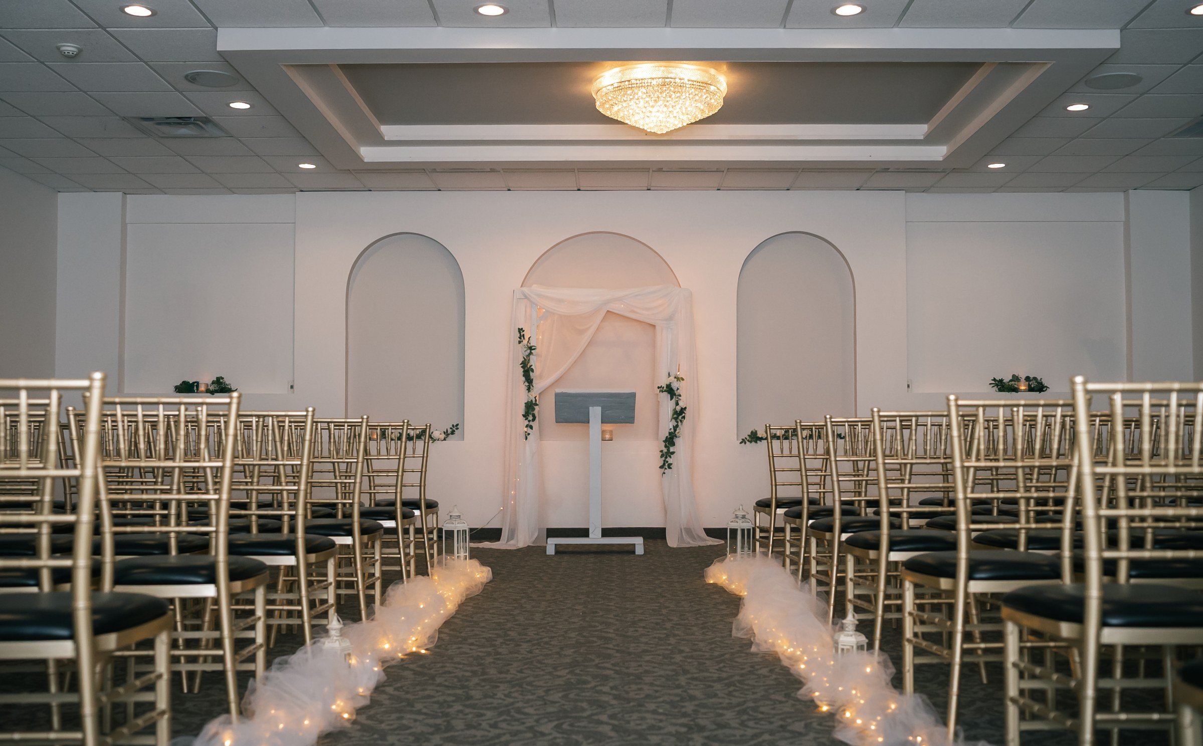 The ceremony room at The Riverdale set before guests arrive: rows of gold chairs, an aisle lined with tulle and lanterns, white arched alcoves at the front and a crystal chandelier overhead