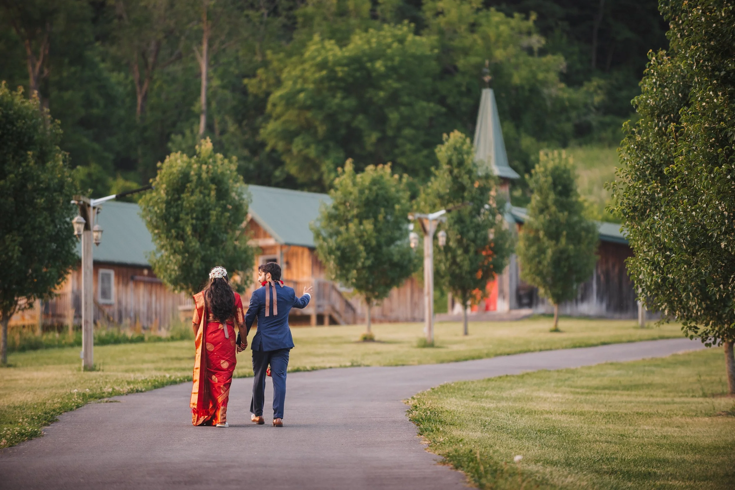 Natalia in red and Ansel in a navy suit walking hand in hand along a path at Wolf Oak Acres, with the barns and a steeple behind them