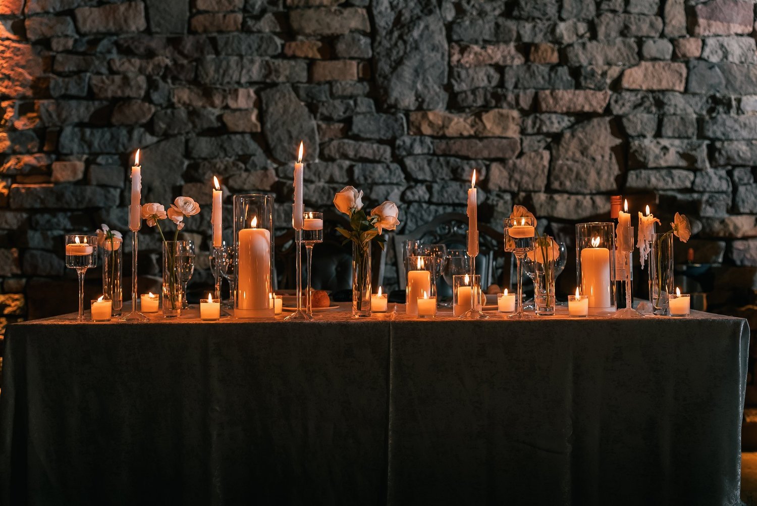 Candles in glass cylinders and flowers arranged across a table beneath The Somerley stone wall.