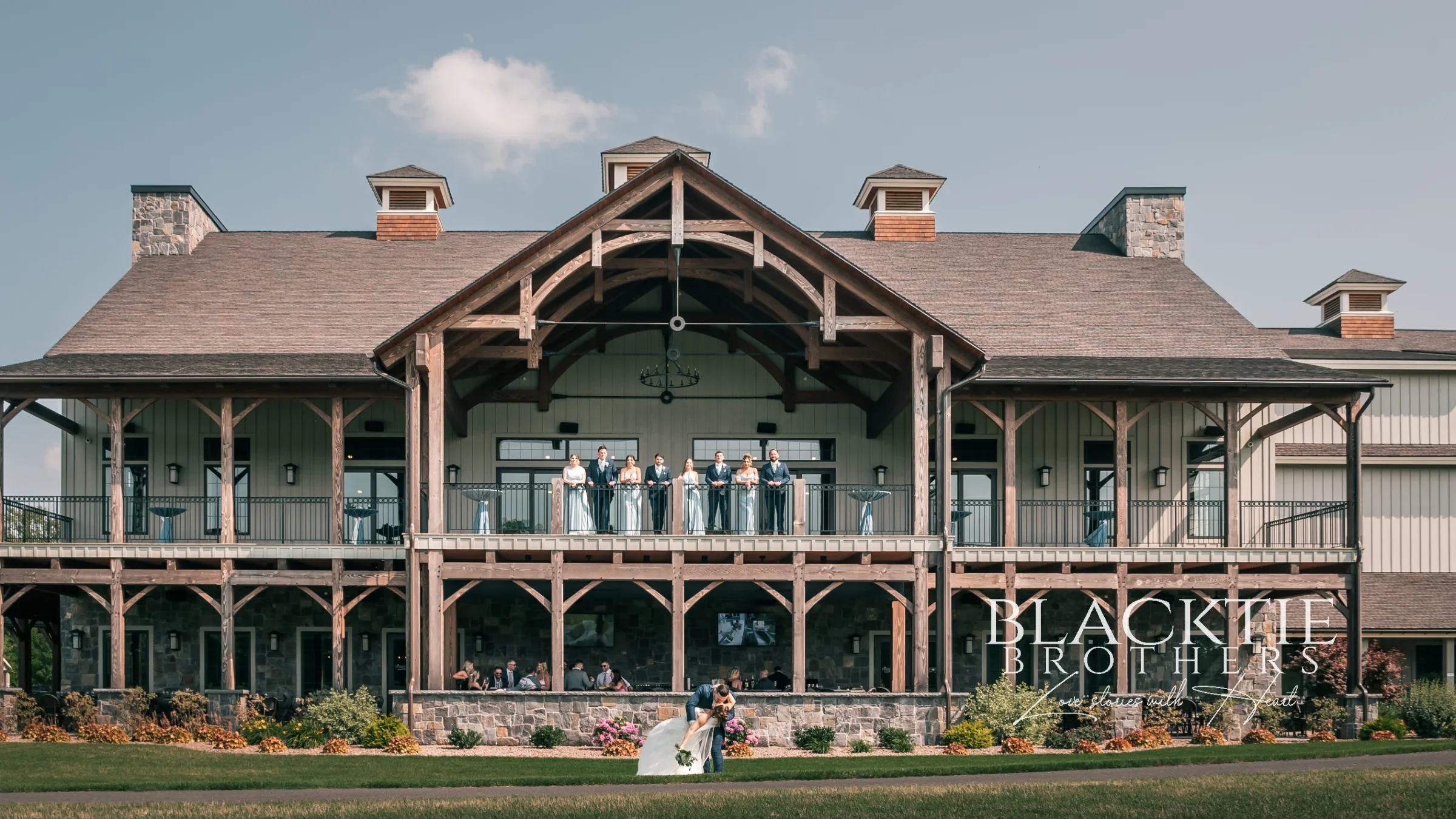 The timber-framed clubhouse at Timber Banks photographed straight on from the lawn under a blue sky, Taylor and Jake's wedding party lined along the upper covered porch beneath the central gable and the couple standing together on the grass below