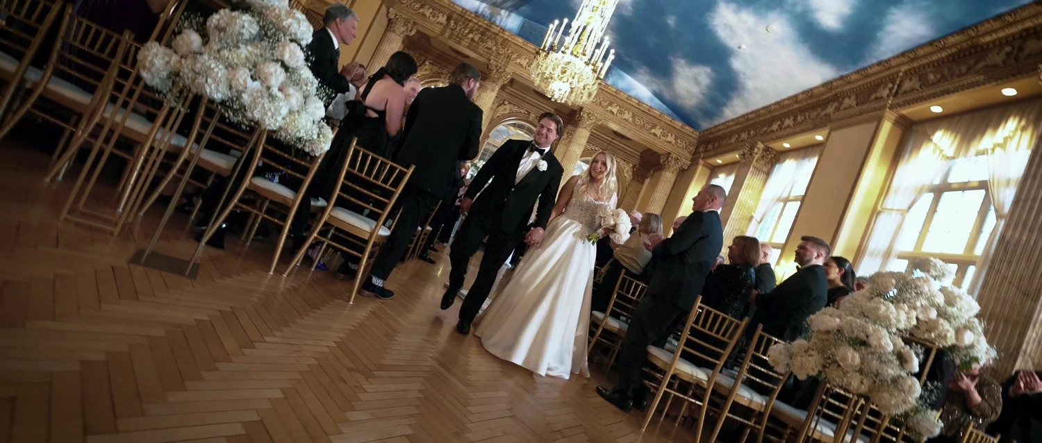 Catie walks into a wedding ceremony in a gold-trimmed room with chandeliers and a blue sky ceiling