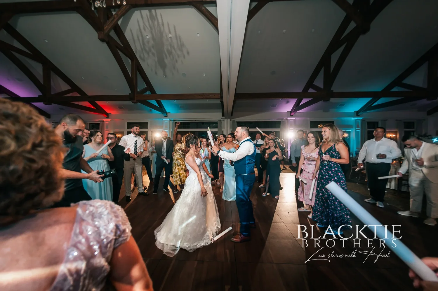 The dance floor at Taylor and Jake's reception seen from above the crowd, guests with light-up foam sticks under the timber trusses and colored uplighting
