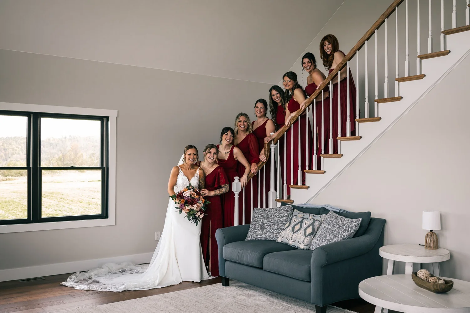 Heather at the foot of a white staircase while her bridesmaids in deep red gowns line the steps above her
