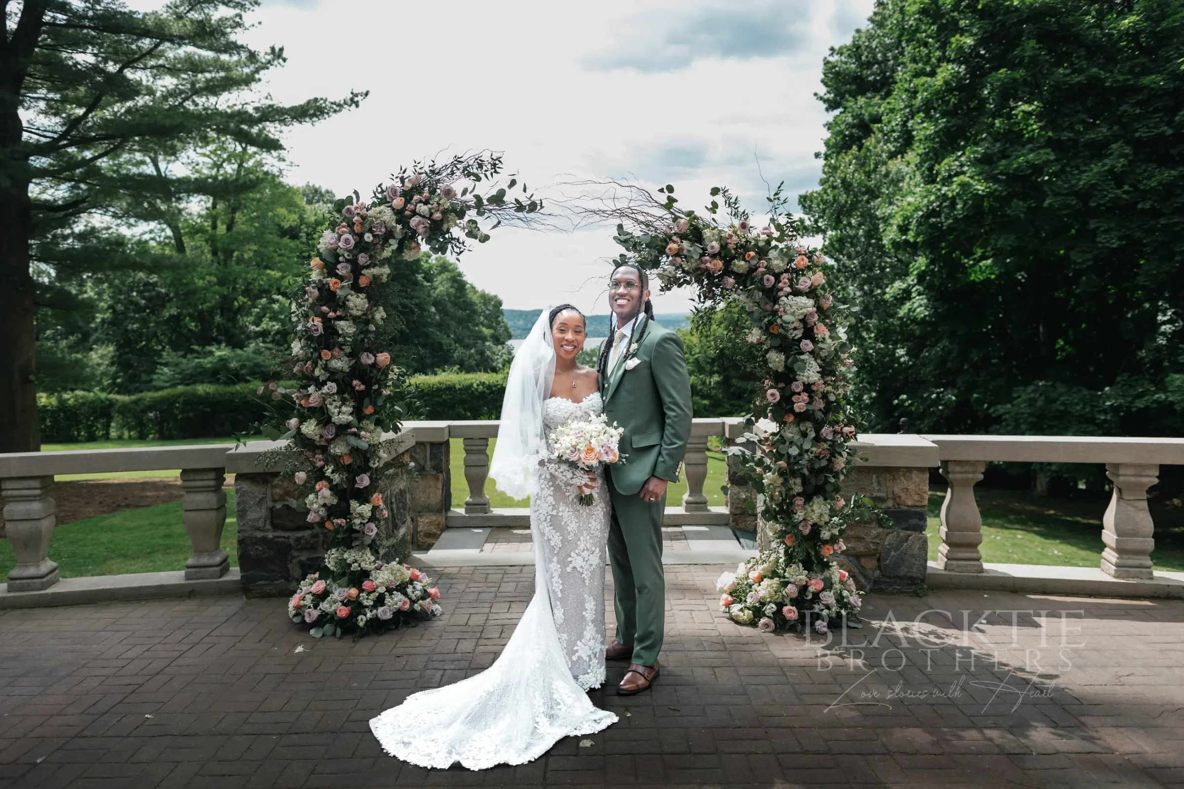 The bride in a lace gown and veil and the groom in a sage-green suit standing between two floral arches on the stone terrace at Tappan Hill Mansion, with the balustrade, trees and the Hudson River behind them