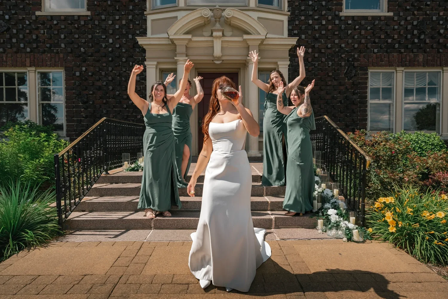 Danielle on the front walk of the mansion at Traditions at the Glen with her bridesmaids in green cheering on the steps behind her
