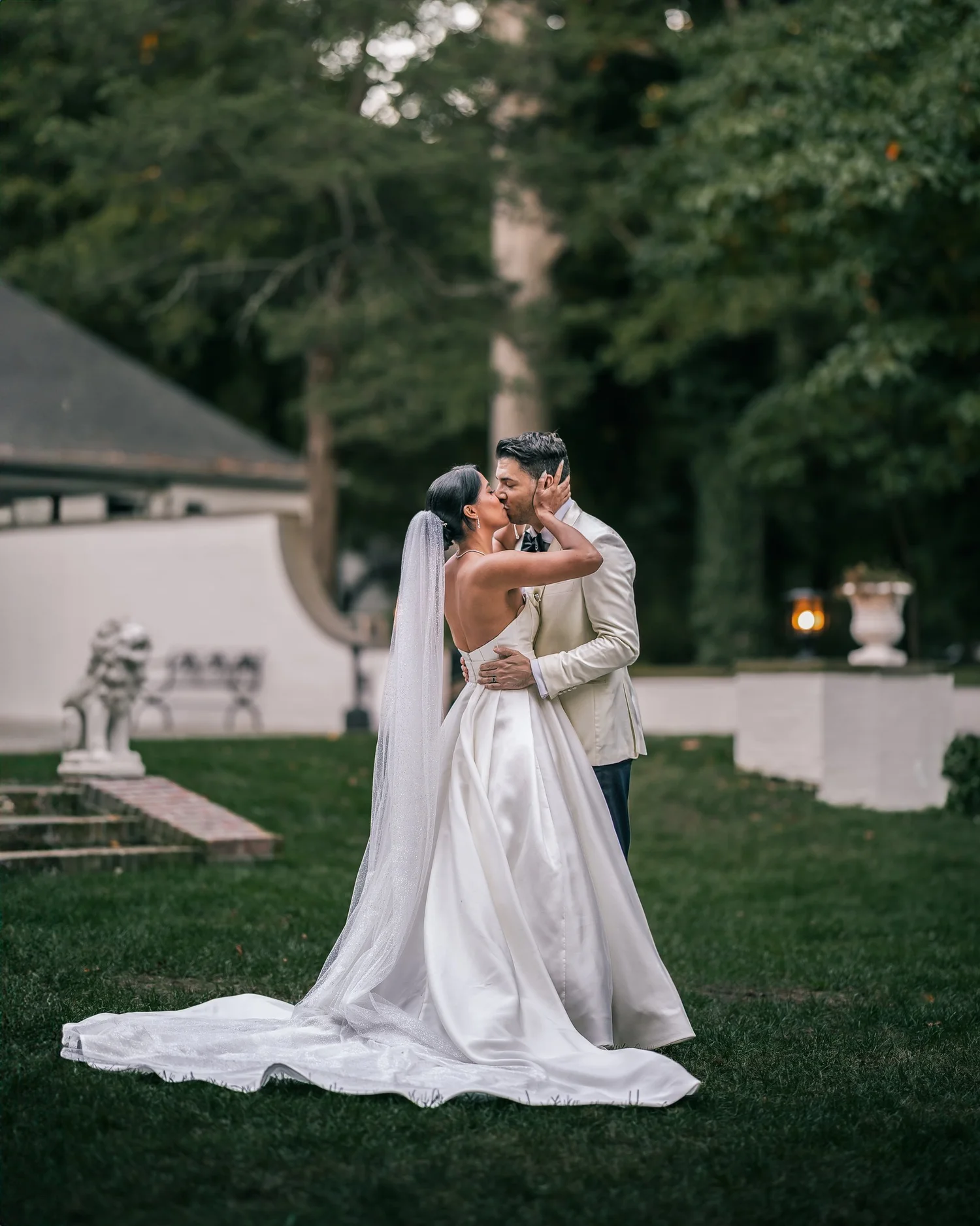 Patricia and Joshua kiss on a lawn, her long veil and train spread across the grass, a stone lion statue behind them.