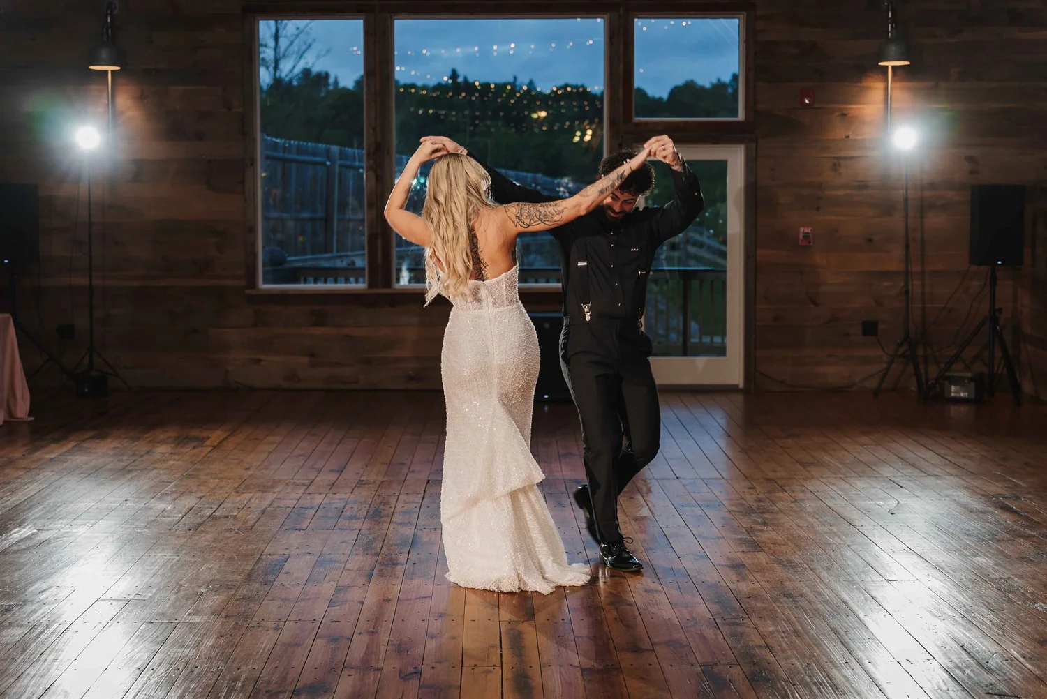 Kari and Josh turn together on the wooden dance floor in front of tall windows at dusk.