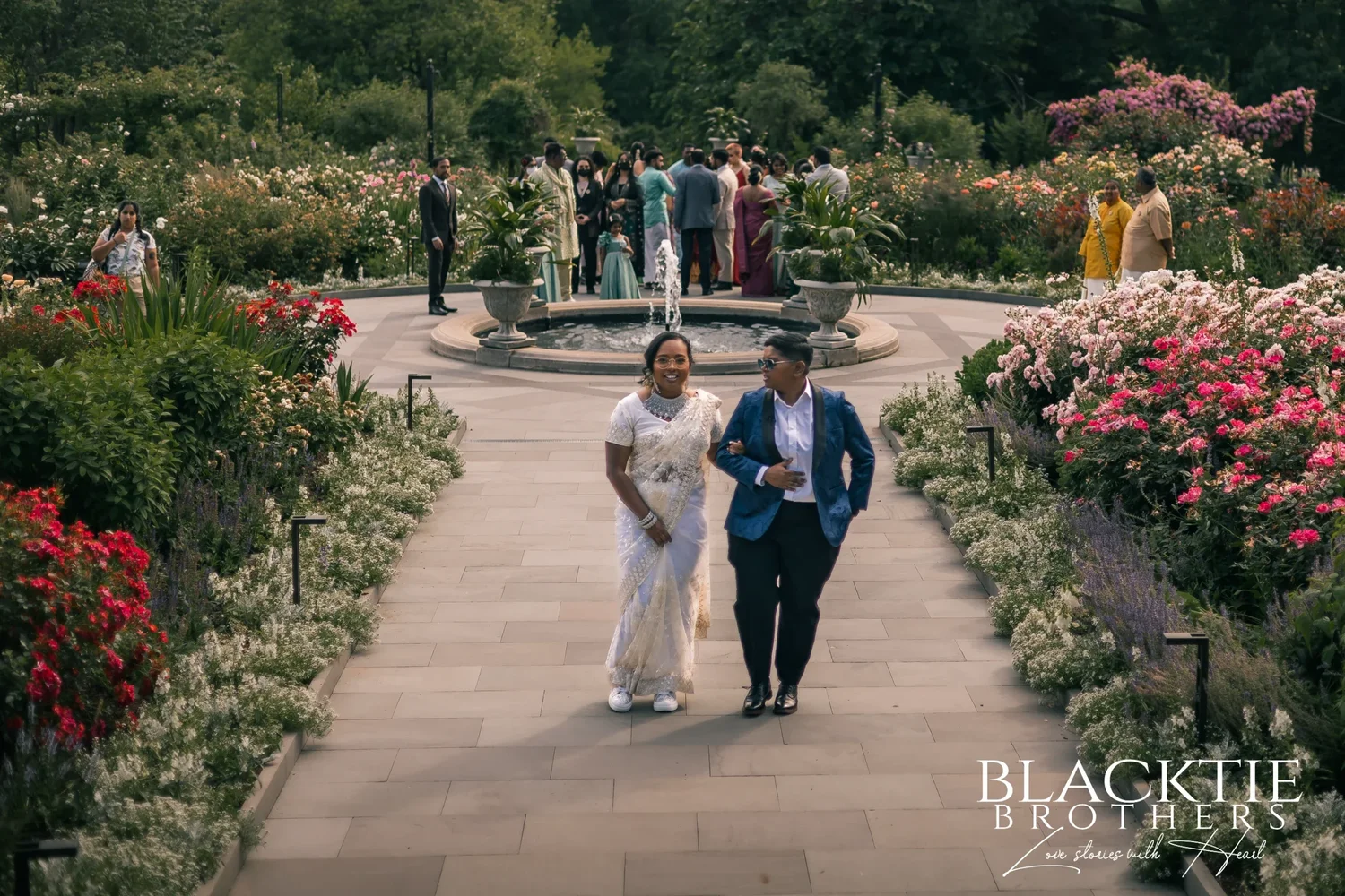 Hima and Alina walking arm in arm down the stone path of the Rose Garden at Morris Arboretum, flower beds on both sides and the round fountain and their guests behind them