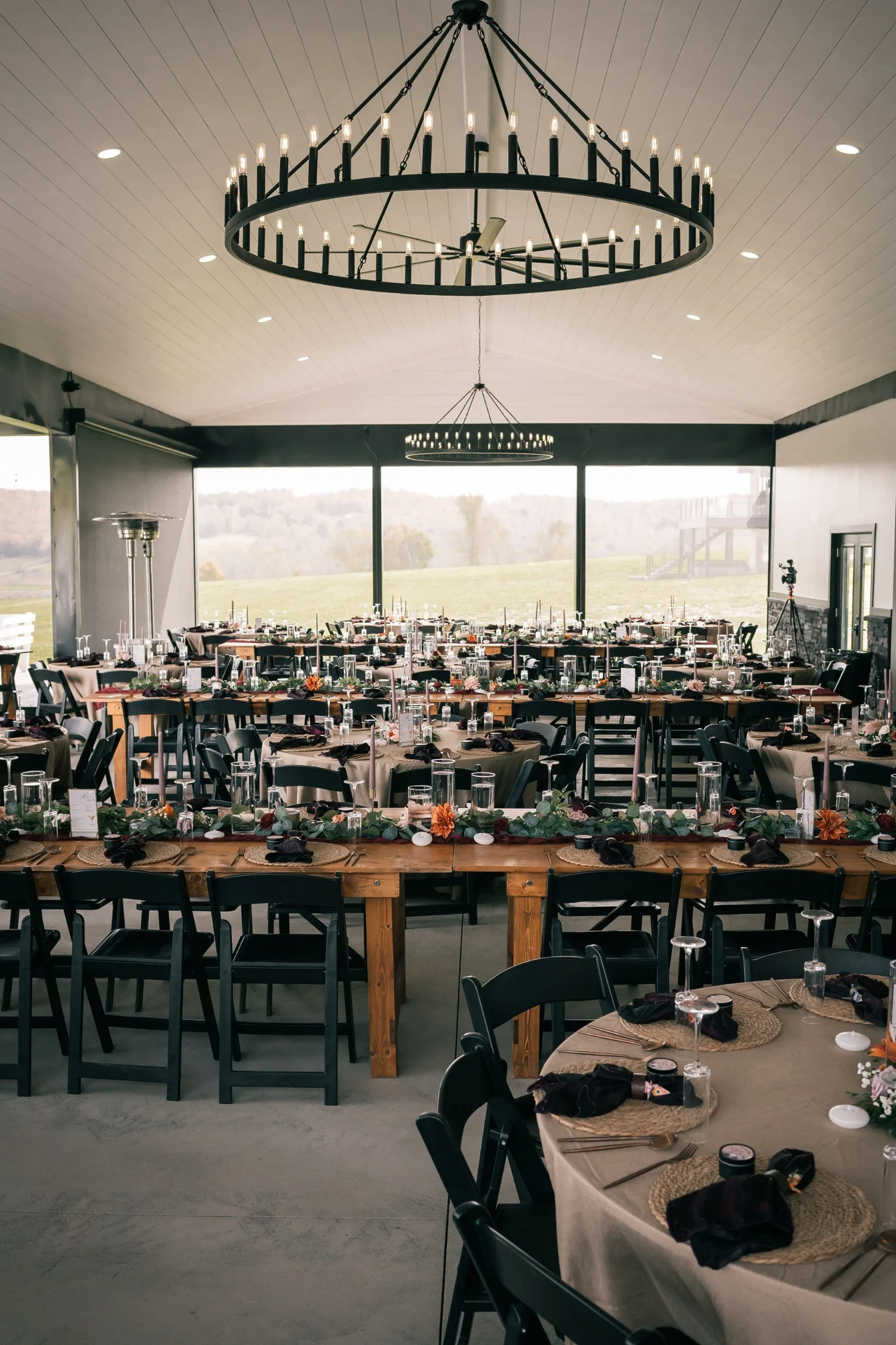 The pavilion before guests arrive, a round iron chandelier above long tables and tall windows facing the pond