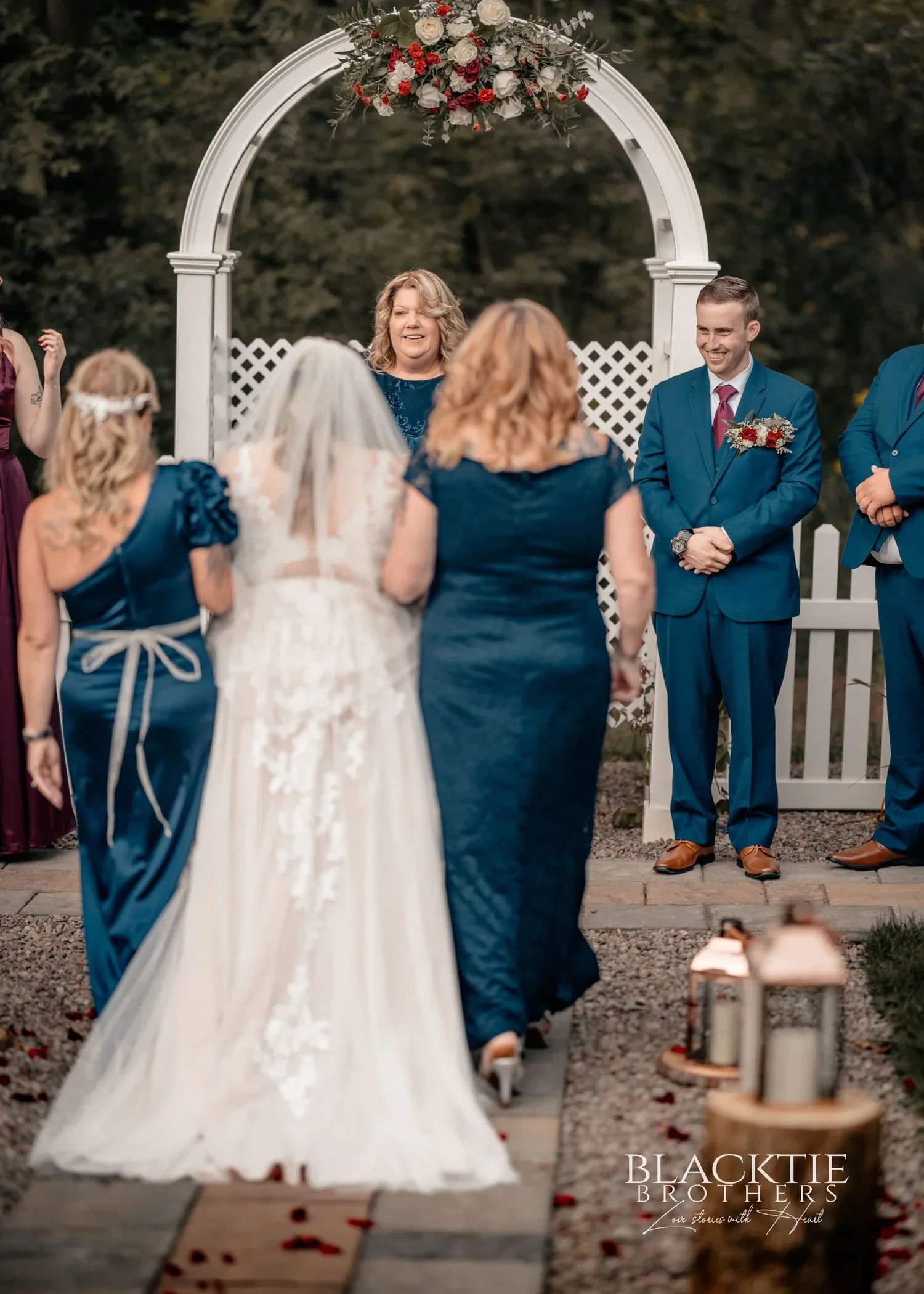 Seen from behind, Jennifer and a woman in navy walk the last steps of the aisle toward the white arbor where Jared waits with his groomsmen