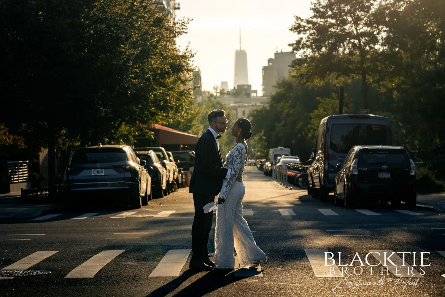 Ciara and Drew face to face in the middle of a sunlit Brooklyn crosswalk, parked cars and trees lining the street and One World Trade Center rising at the far end
