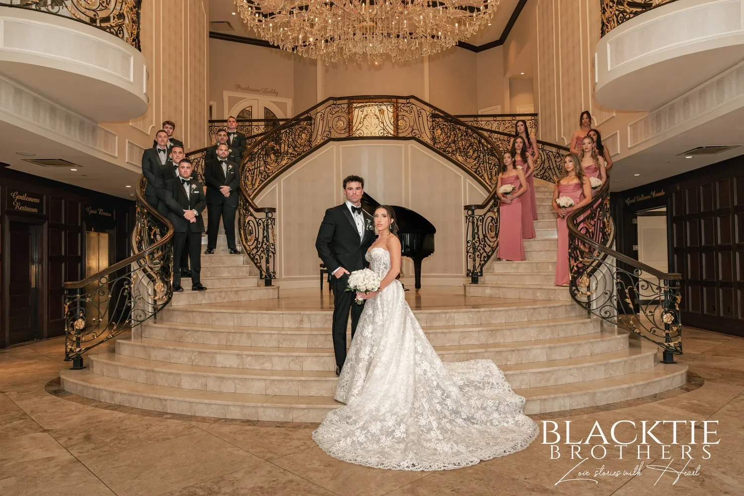 Wide view of the entry hall at The Surf Club on the Sound: a curving marble double staircase with wrought-iron railings under a crystal chandelier, Leah and Nicholas at the foot of the stairs and the wedding party in black tie and dusty rose on both sides