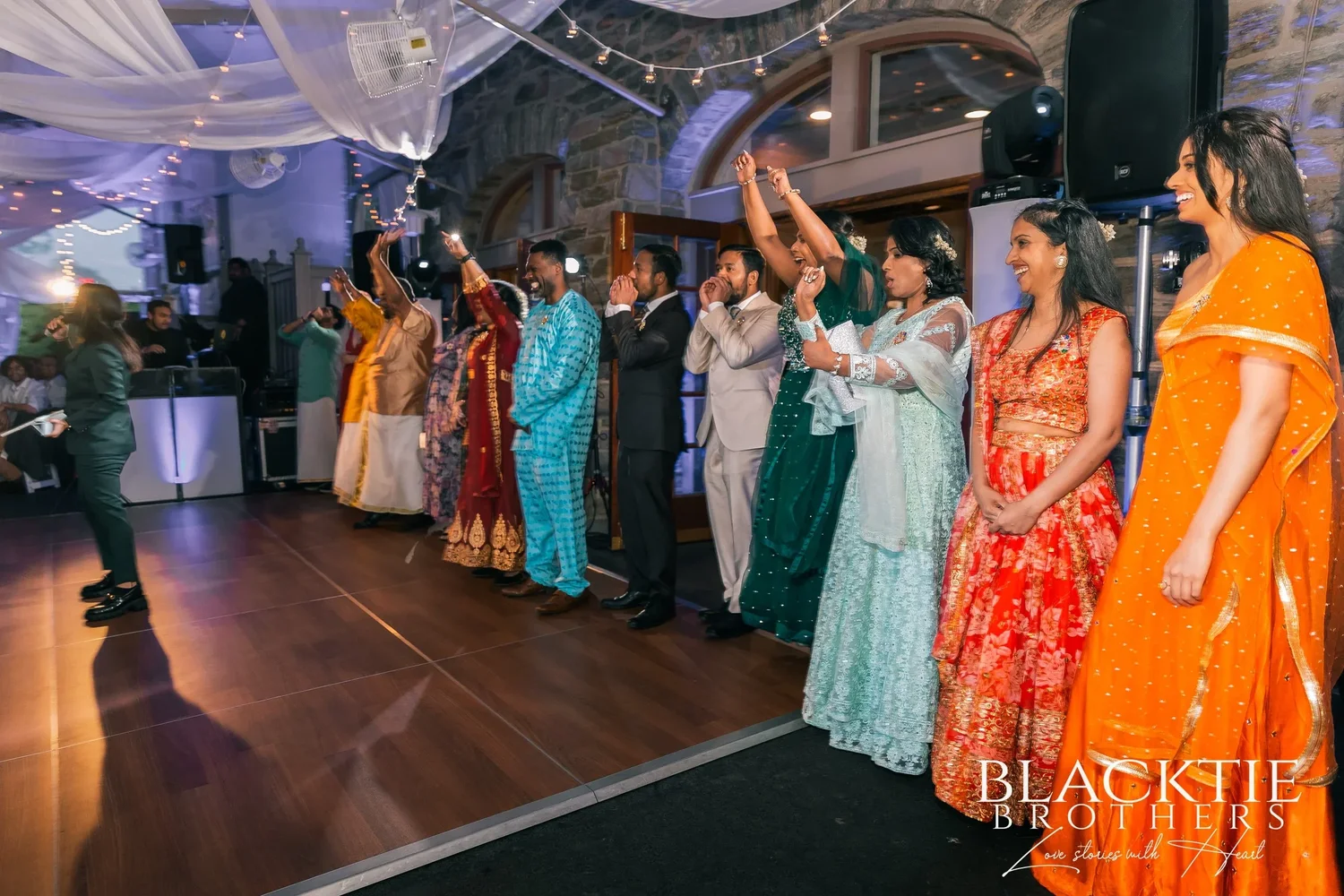 Guests in bright saris and suits lining the dance floor in the reception tent, the stone wall and arched windows of the carriage house behind them