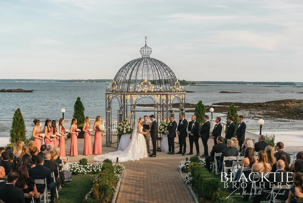 Wedding ceremony at The Surf Club on the Sound in New Rochelle, New York: the couple under the domed iron gazebo at the end of a brick aisle, wedding party on both sides and Long Island Sound behind them