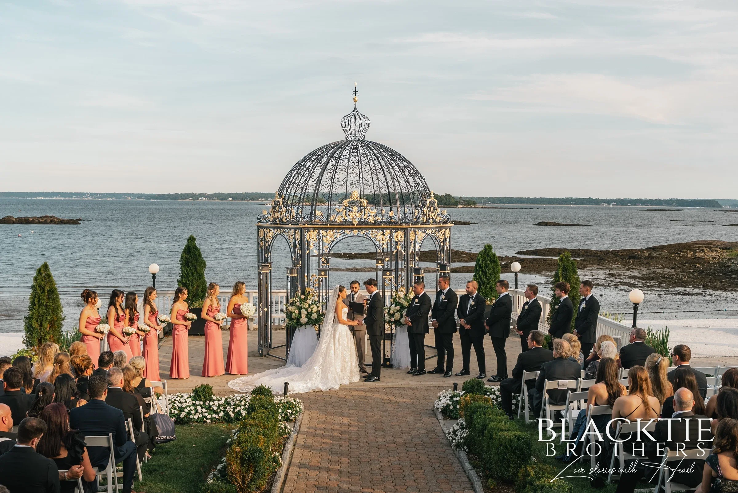 The ceremony at The Surf Club on the Sound: Leah and Nicholas facing each other under the ornate domed iron gazebo at the end of a brick aisle, bridesmaids in dusty rose and groomsmen in black tie on either side, guests seated in the foreground and Long Island Sound behind them