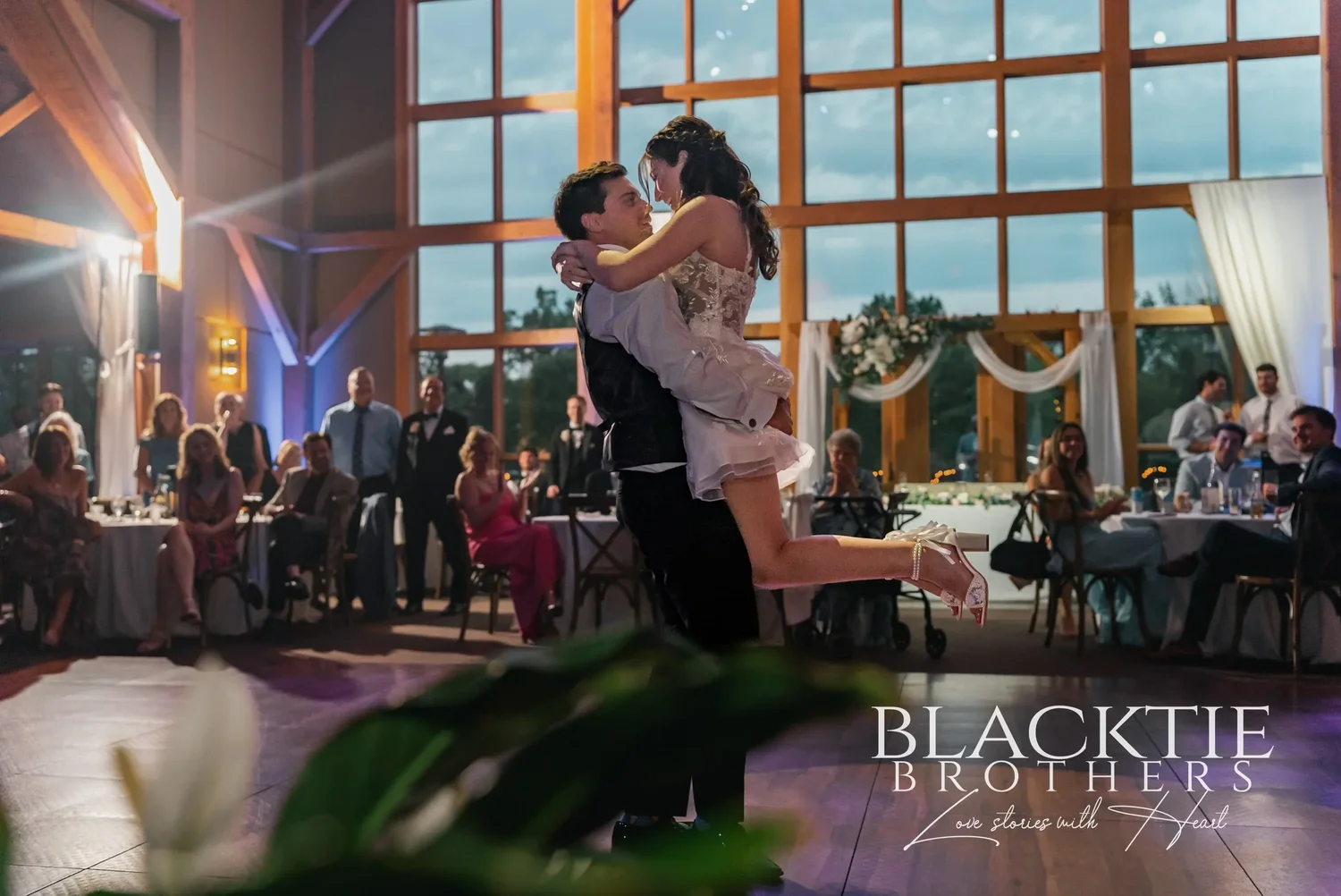 A groom lifting his bride on the dance floor at Allyn Lodge at dusk, with the window wall glowing behind them.