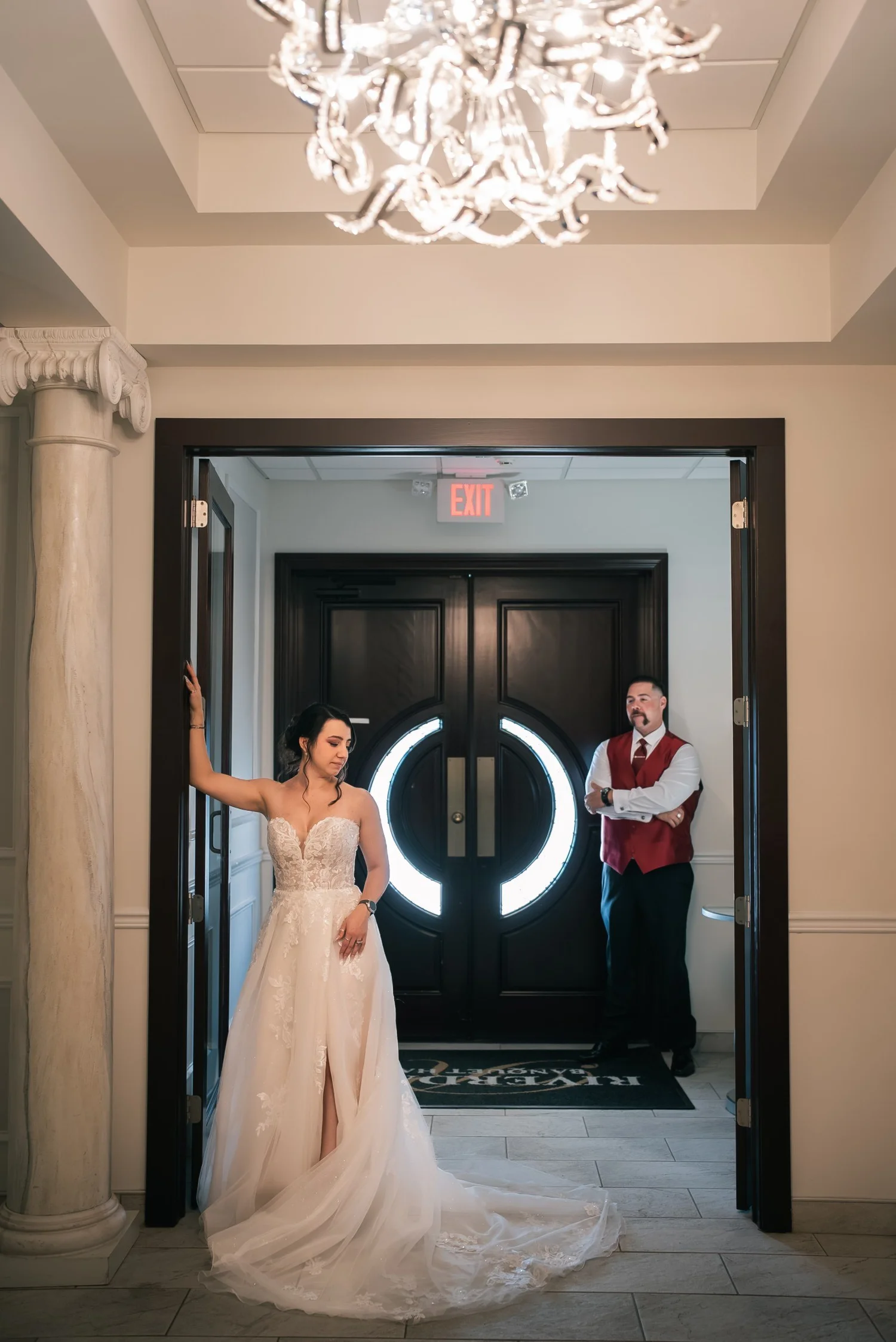 Laura standing in a doorway under a crystal chandelier, Dante waiting by the dark entry doors behind her