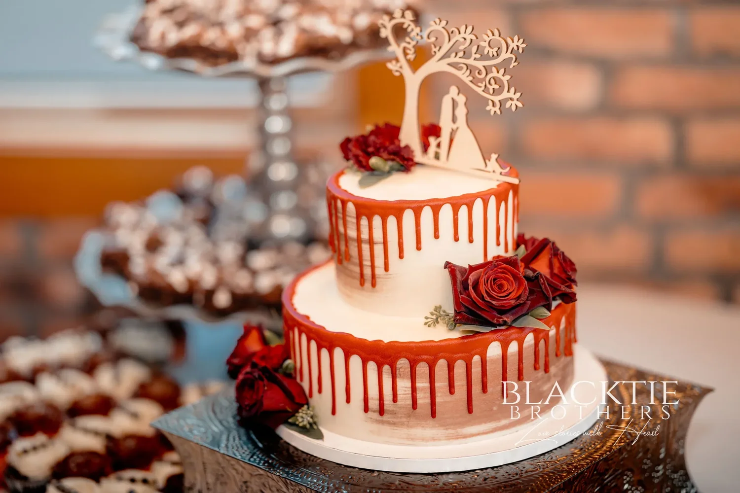 A two-tier white wedding cake with red drip icing, deep red roses and a wooden tree cake topper, cupcakes beside it on a brick-walled table