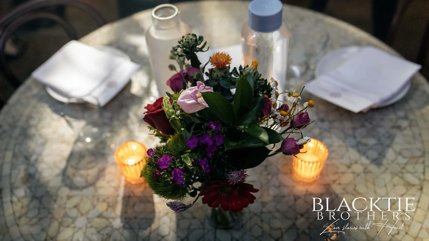A mosaic tile table at Fandi Mata set with a low arrangement of red, pink, purple and orange flowers, two glowing candle holders and folded white napkins