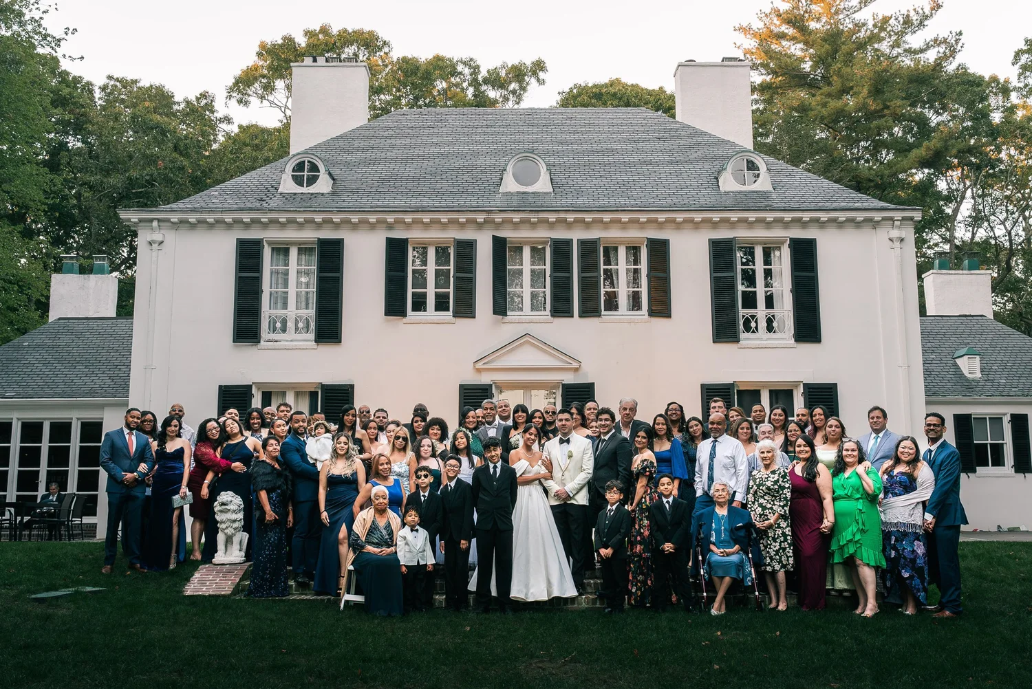 A large group of family and friends in formal clothes gathered around Patricia and Joshua in front of a white house with black shutters.
