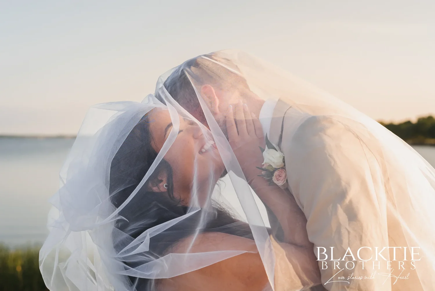 Carolina and Josh close together under her veil in warm evening light by the water