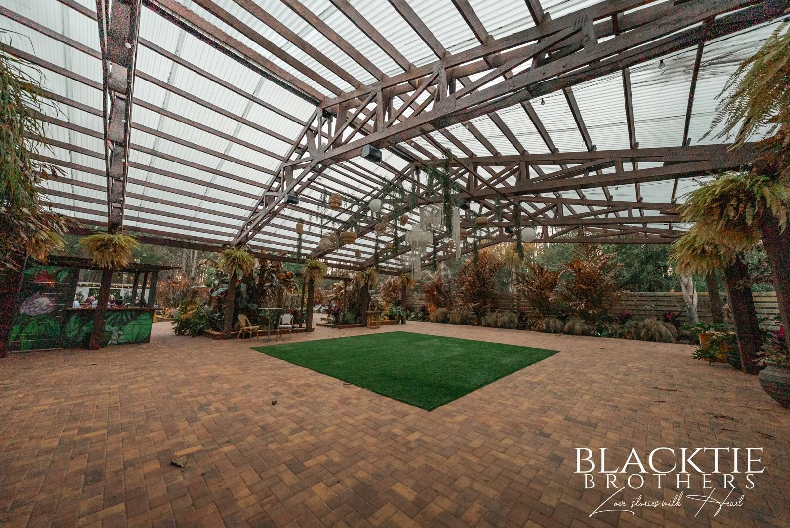 An open-sided covered event space has a pitched translucent roof, hanging greenery and a central green dance area.