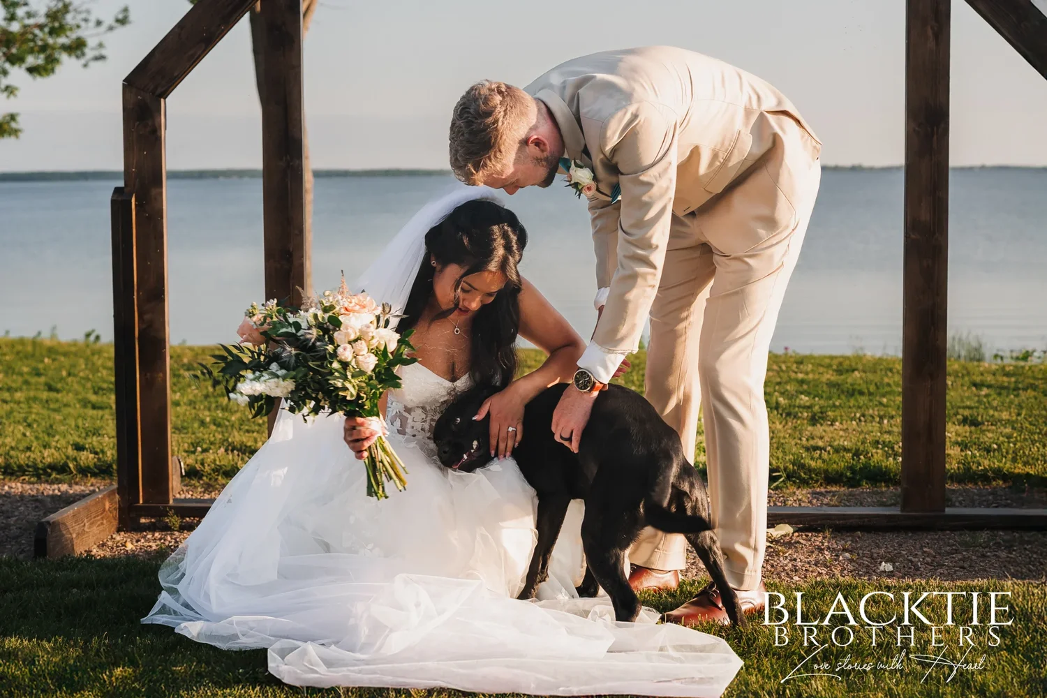 Carolina crouching to hug a black dog while Josh leans down beside them under the wooden arch, the water behind them