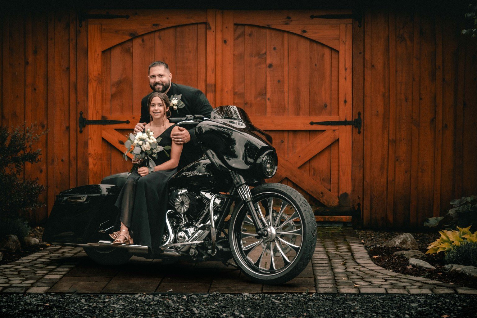 Louie poses with a wedding guest seated on a black motorcycle in front of the wooden barn doors.