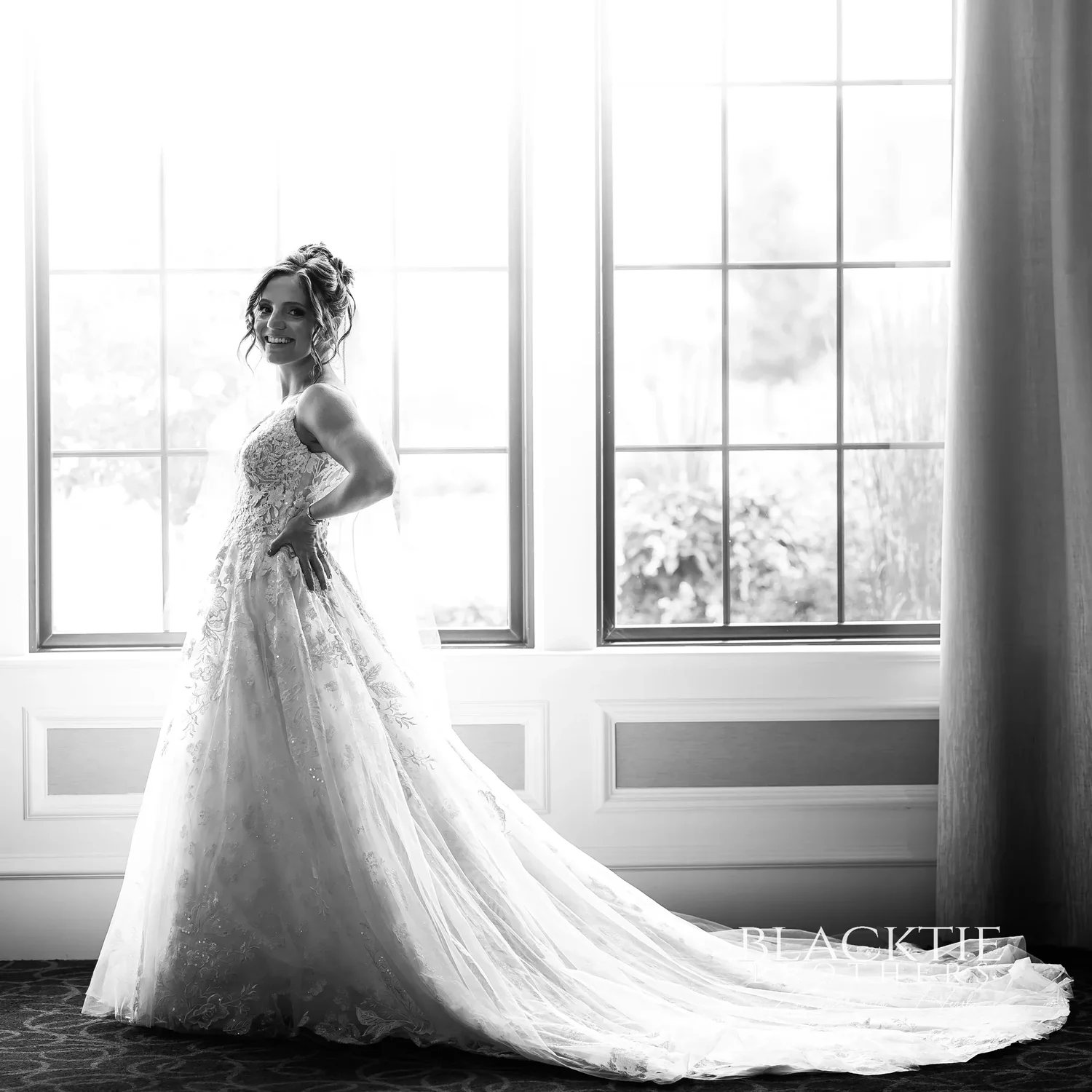 Black and white portrait of Taylor in her wedding dress standing in front of the tall suite windows, backlit, her train spread across the carpet