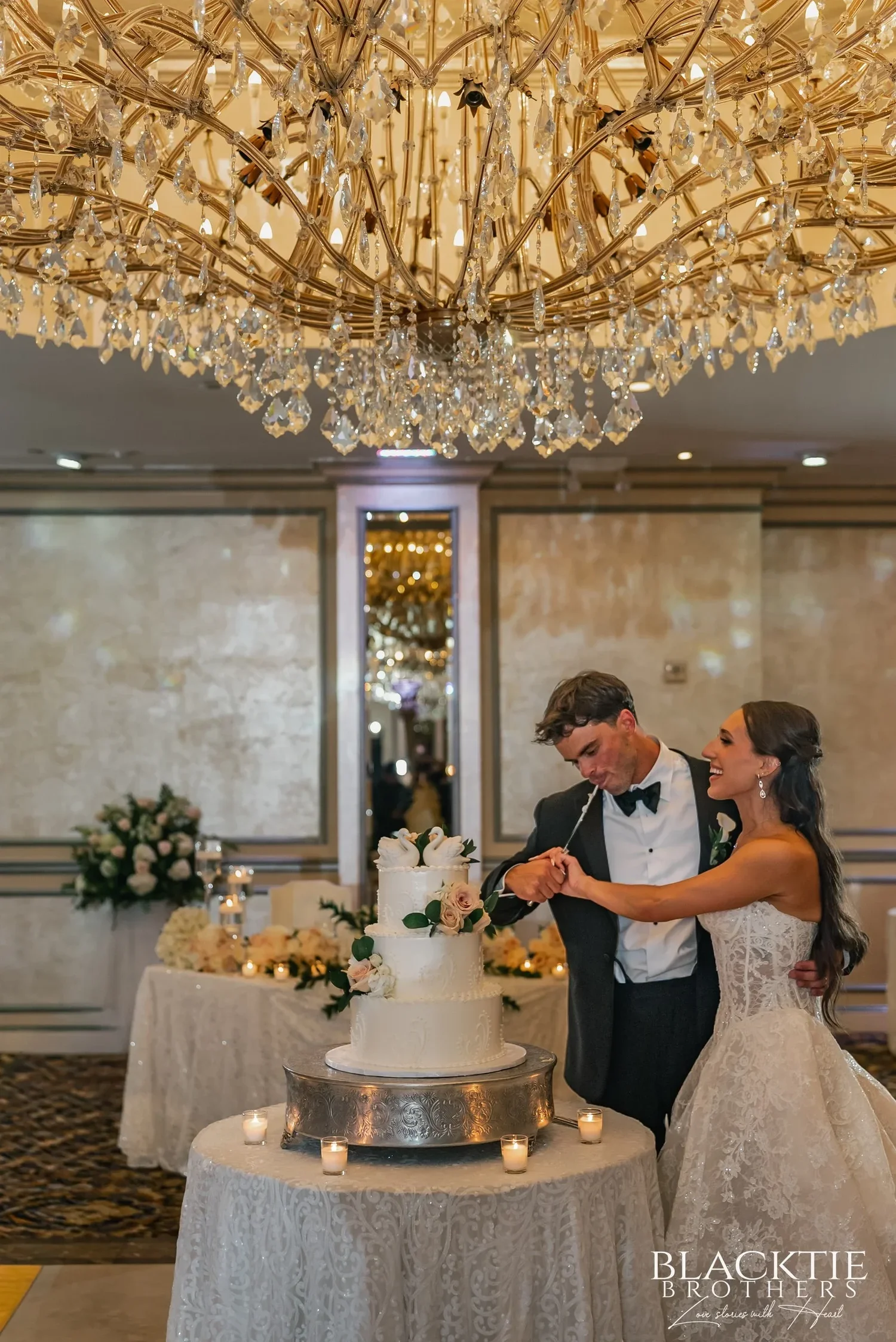 Leah and Nicholas cutting a tiered white wedding cake together beneath a large gold and crystal chandelier