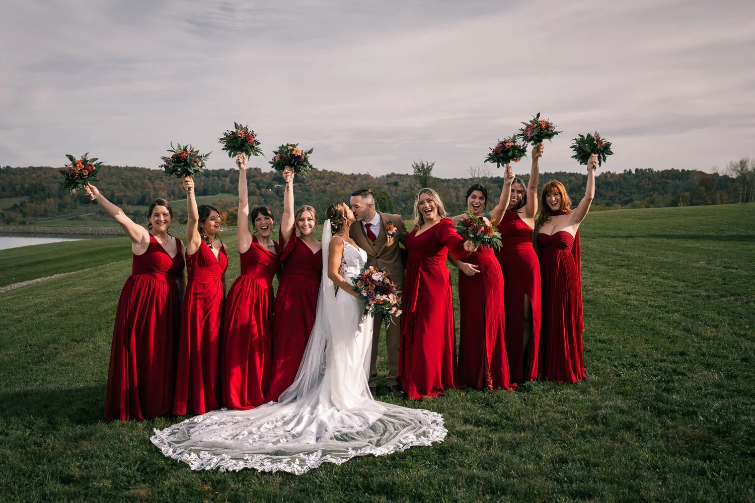 Heather and her bridesmaids in red gowns raising their bouquets on the lawn