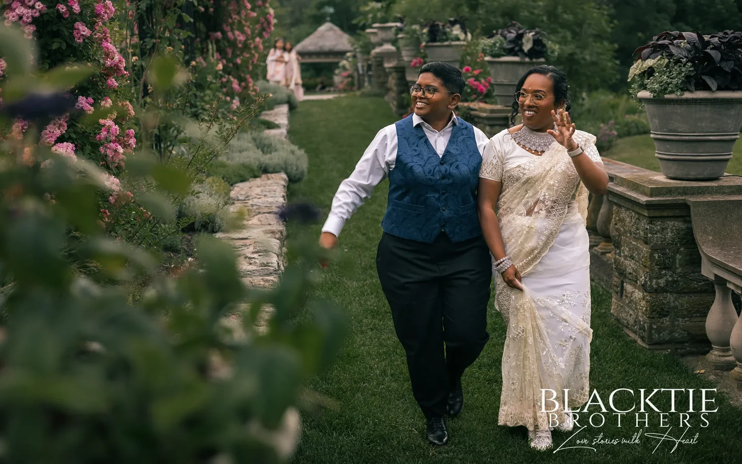 Hima and Alina walking and laughing along a grassy garden path beside a stone balustrade, pink roses and a small gazebo behind them