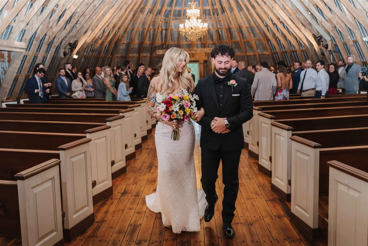 Kari and Josh walk arm in arm between the white pews as guests stand behind them.