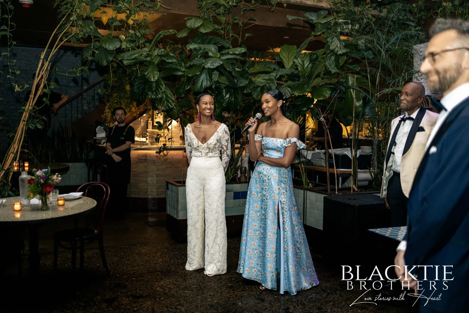 Ciara standing beside a member of the wedding party in a pale blue gown who gives a toast into a microphone, surrounded by plants and candlelit tables