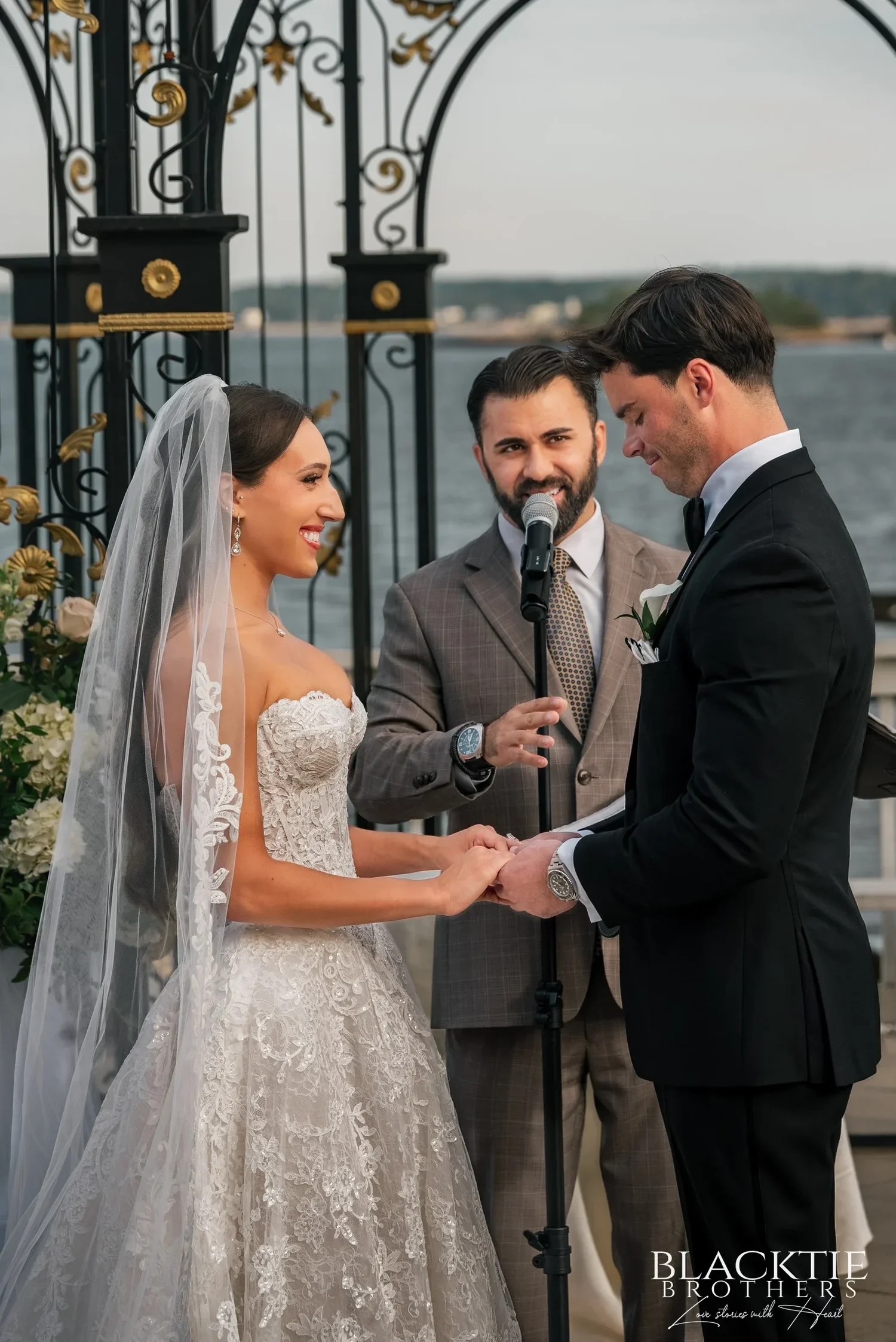 Leah and Nicholas holding hands and laughing during the ring exchange under the iron gazebo, the officiant speaking into a microphone beside them and the water behind