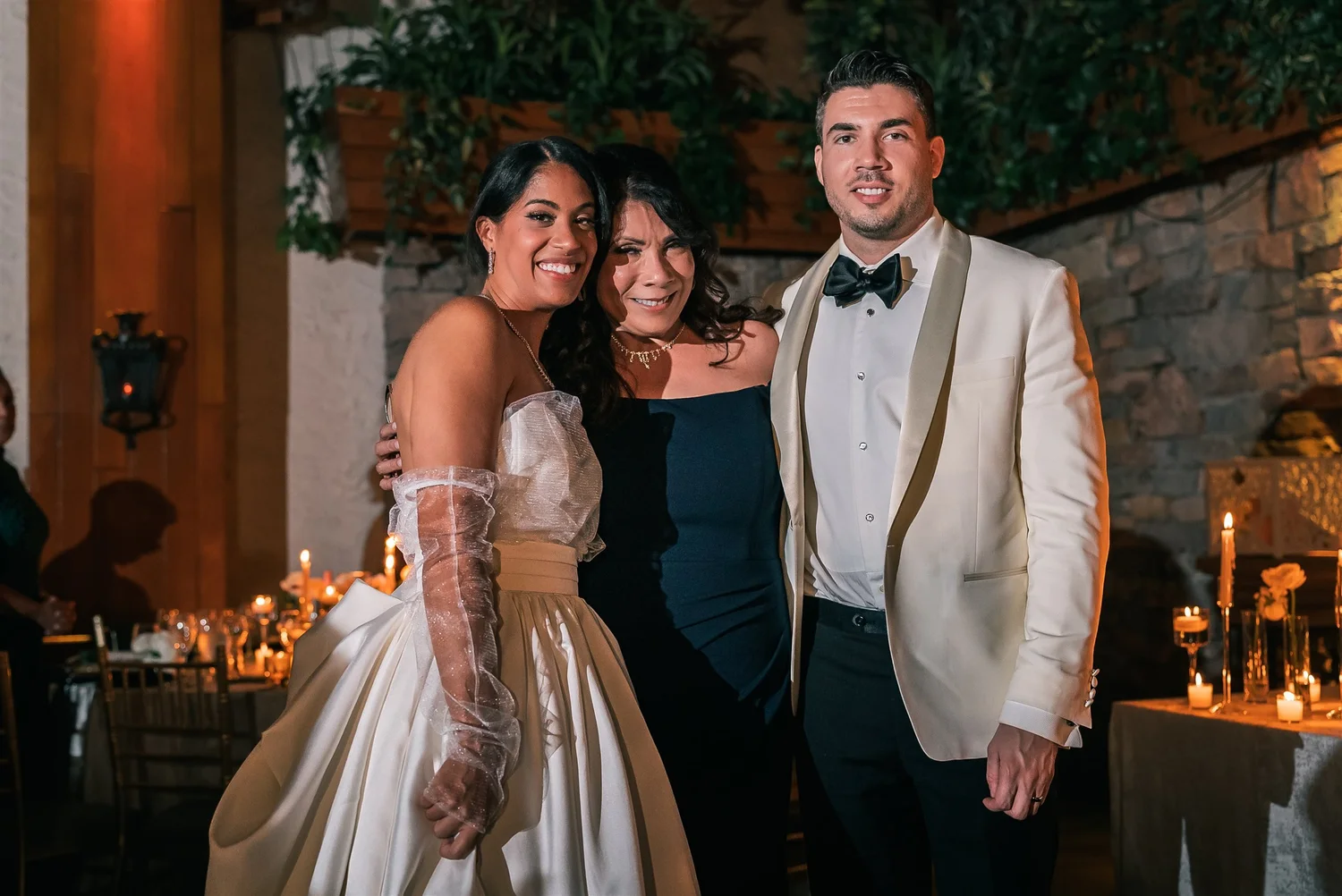 Patricia and Joshua pose with a guest beside the candlelit tables and stone wall at The Somerley.