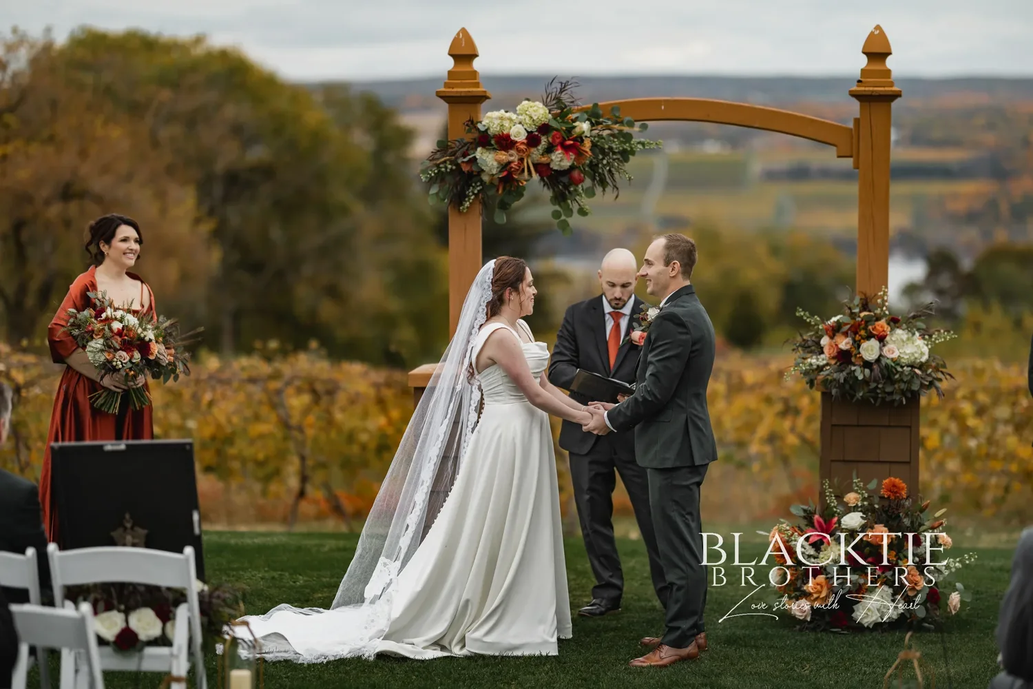 Anya and Gabe holding hands at the wooden arbor during their ceremony at Glenora Wine Cellars, the officiant between them and the vineyard behind