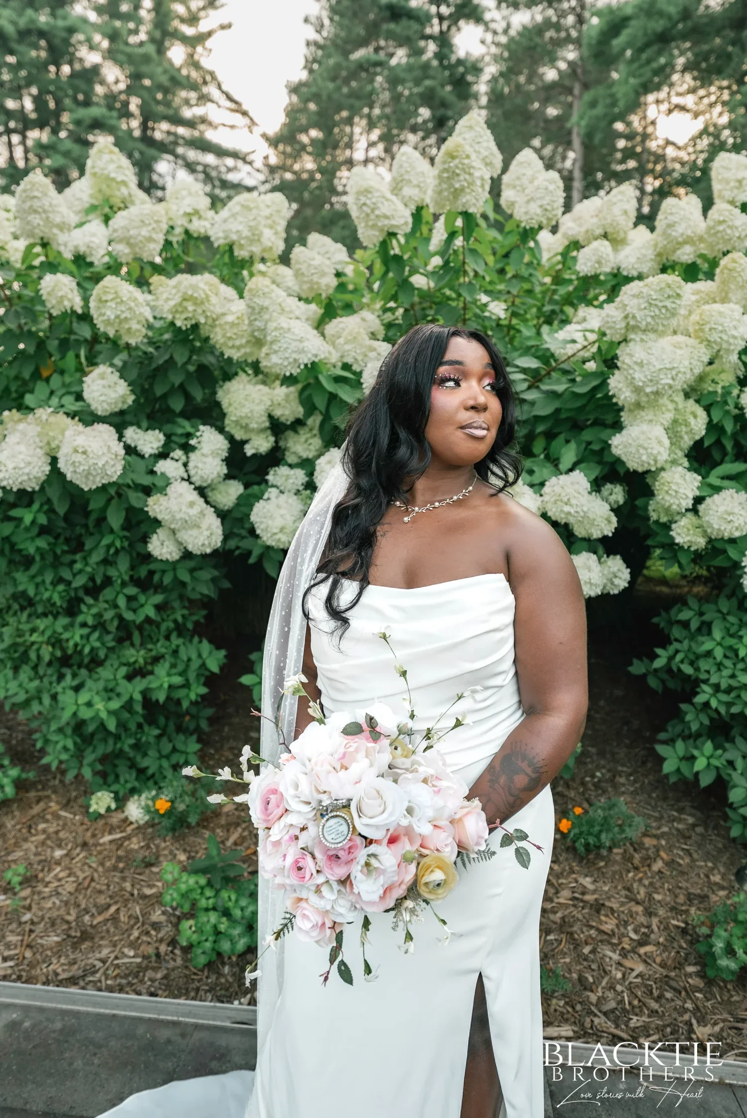 A bride in a strapless satin gown and long veil holding a pink and white bouquet beside the white hydrangeas at Traditions at the Glen