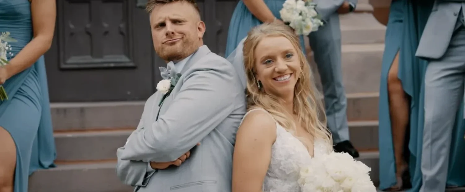 Alex and Jennifer standing back to back on the church steps, he with his arms crossed and a raised eyebrow, she laughing, bridesmaids in dusty blue around them