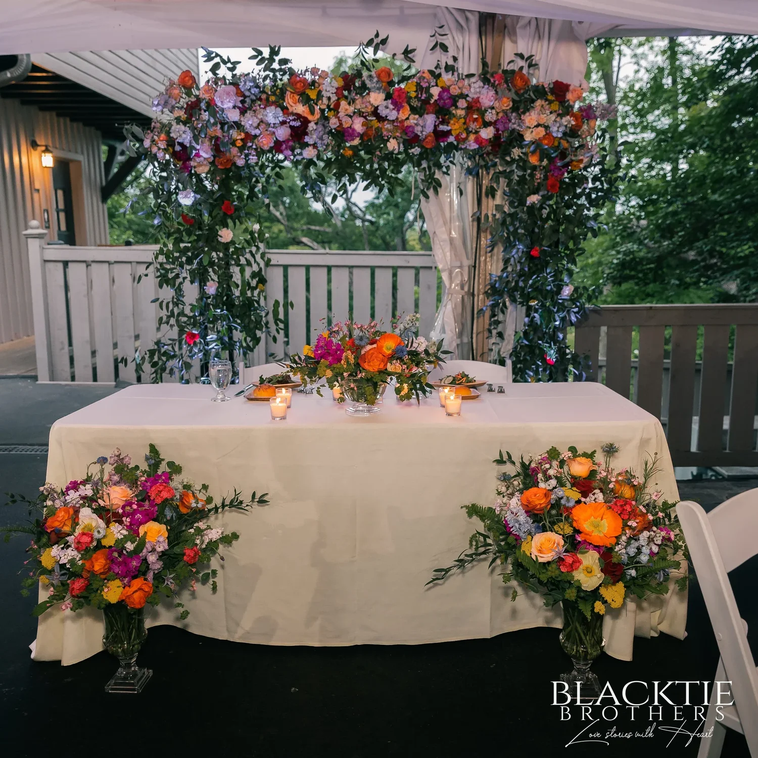 The sweetheart table in the reception tent, set with candles and two bright flower arrangements under an arch of roses and greenery