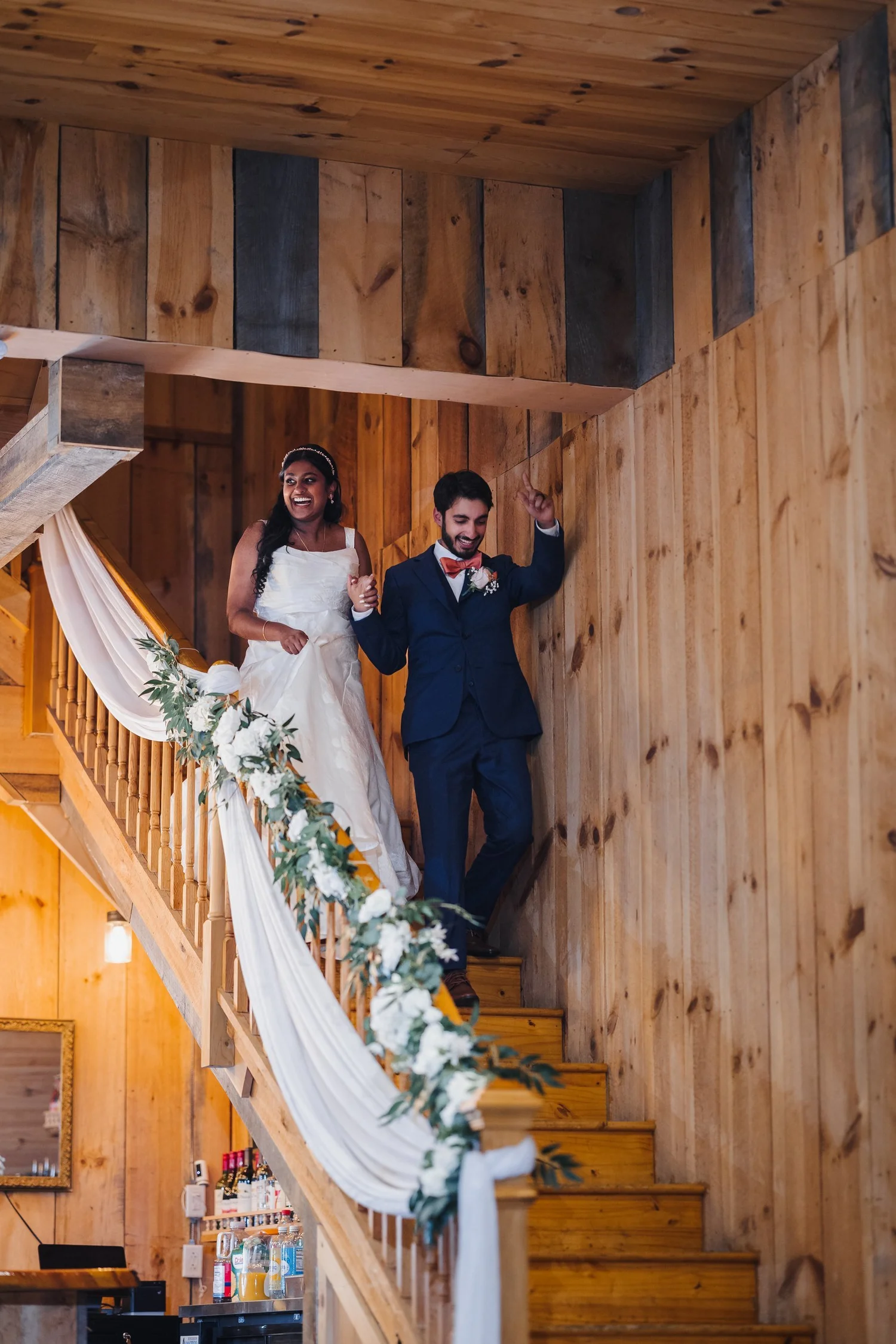 Natalia and Ansel coming down the wooden staircase, its railing draped in white fabric and greenery