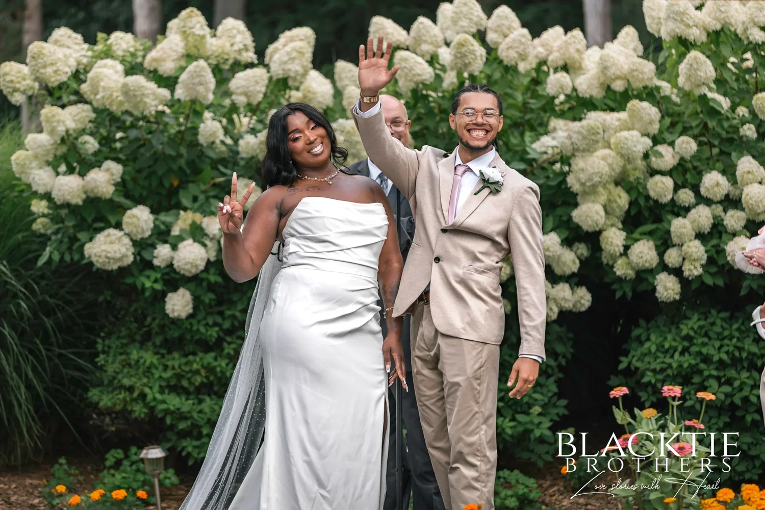 A bride and groom raising their arms in celebration in front of a tall white hydrangea hedge at Traditions at the Glen