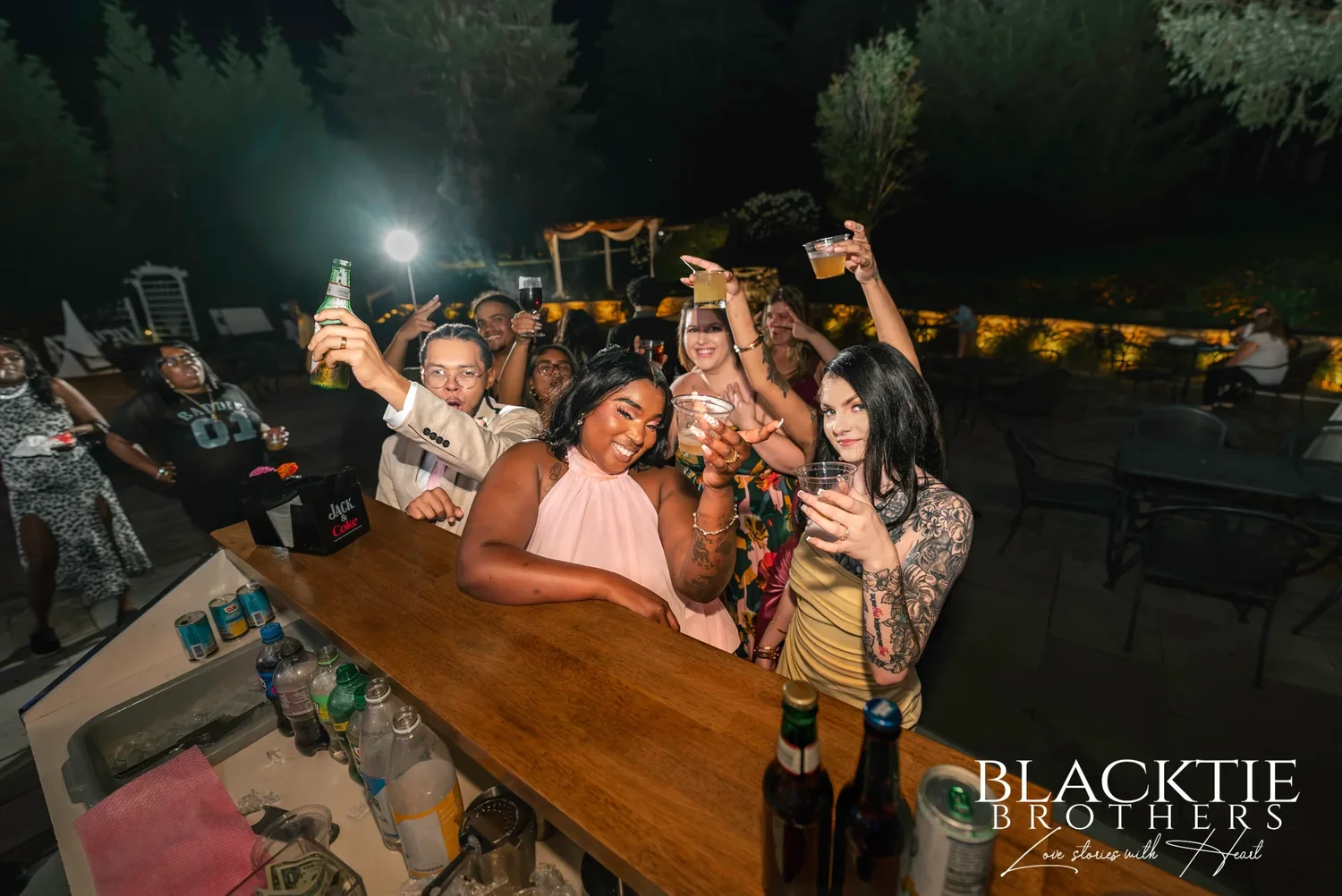 Wedding guests raising their drinks at the outdoor wooden bar at night at Traditions at the Glen
