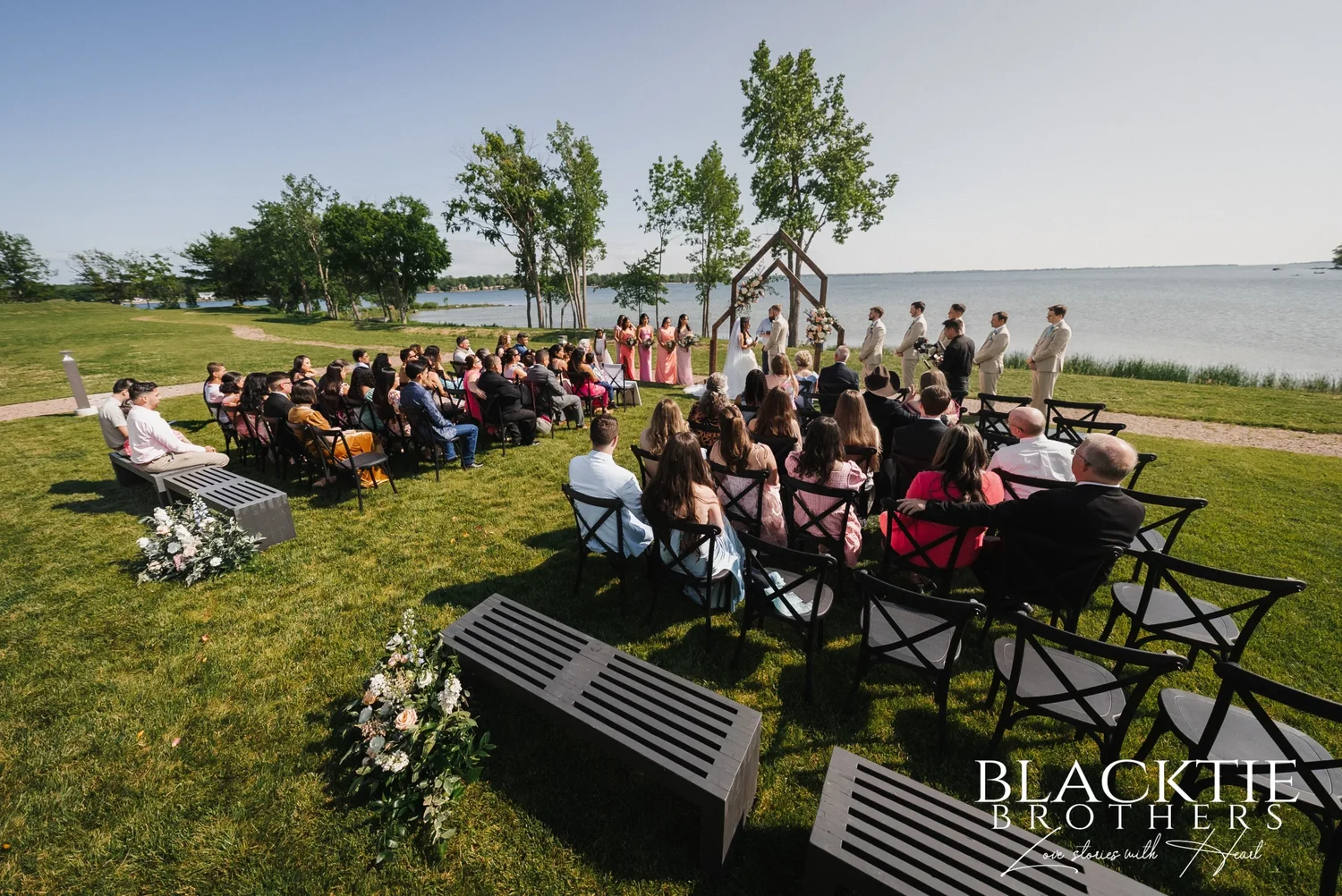 The whole ceremony from the back corner of the lawn: rows of dark chairs filled with guests, the wedding party at the wooden arch and open water reaching the horizon