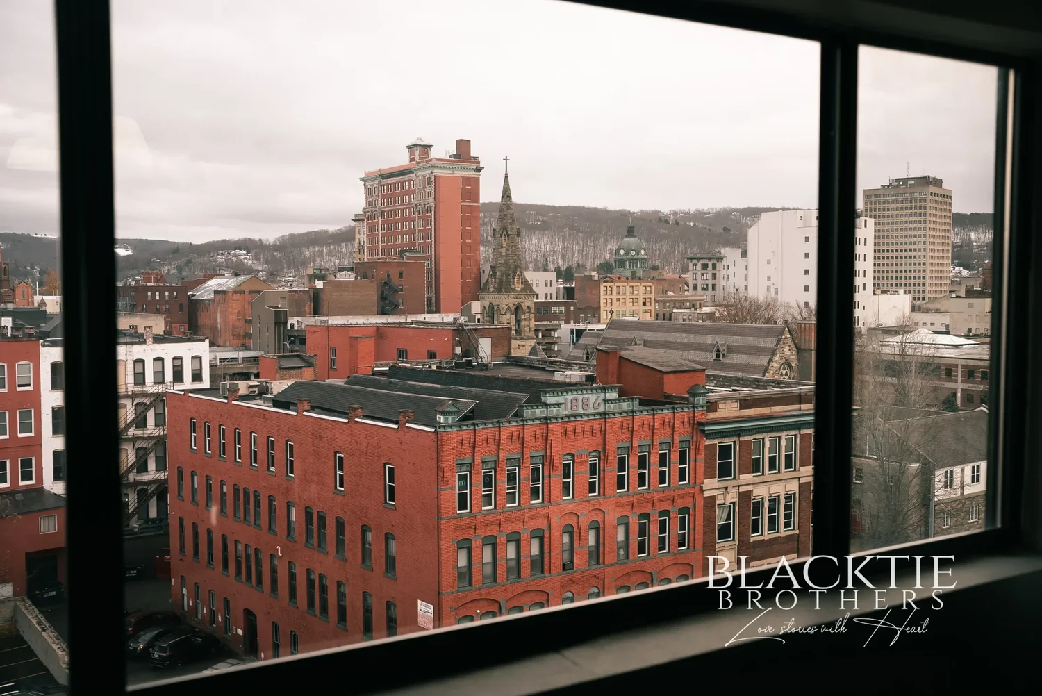 View through a guest room window at the DoubleTree by Hilton Binghamton over red brick downtown buildings, a church spire and the hills under an overcast sky