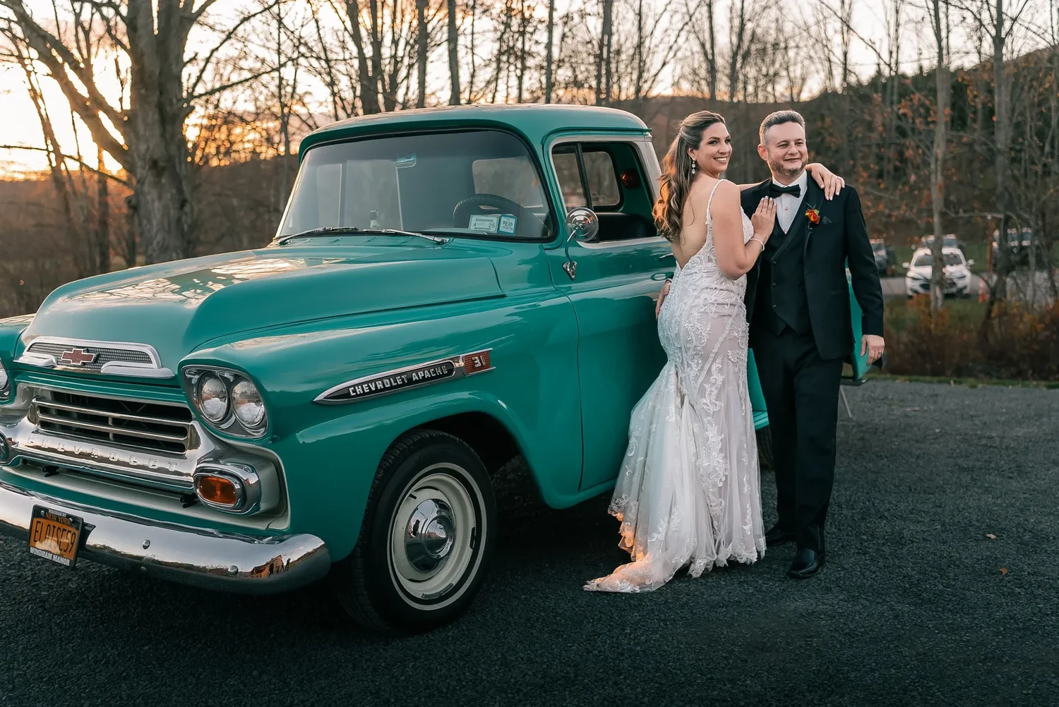 Charissa and Andrew leaning against a vintage teal pickup truck at sunset, the trees bare and golden behind them