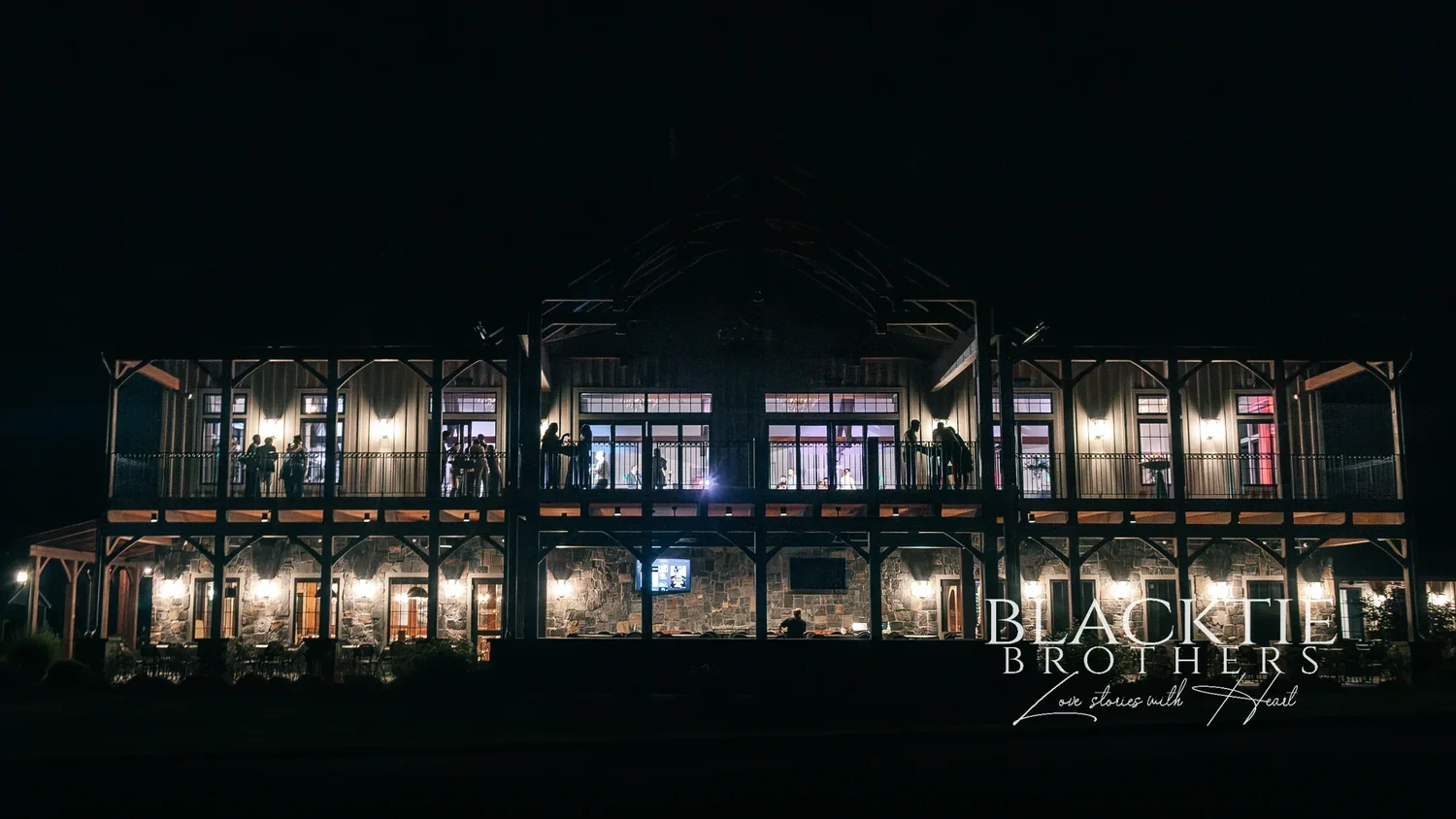 The Timber Banks clubhouse at night photographed straight on, both levels of covered porches glowing with warm light and guests visible on the upper porch
