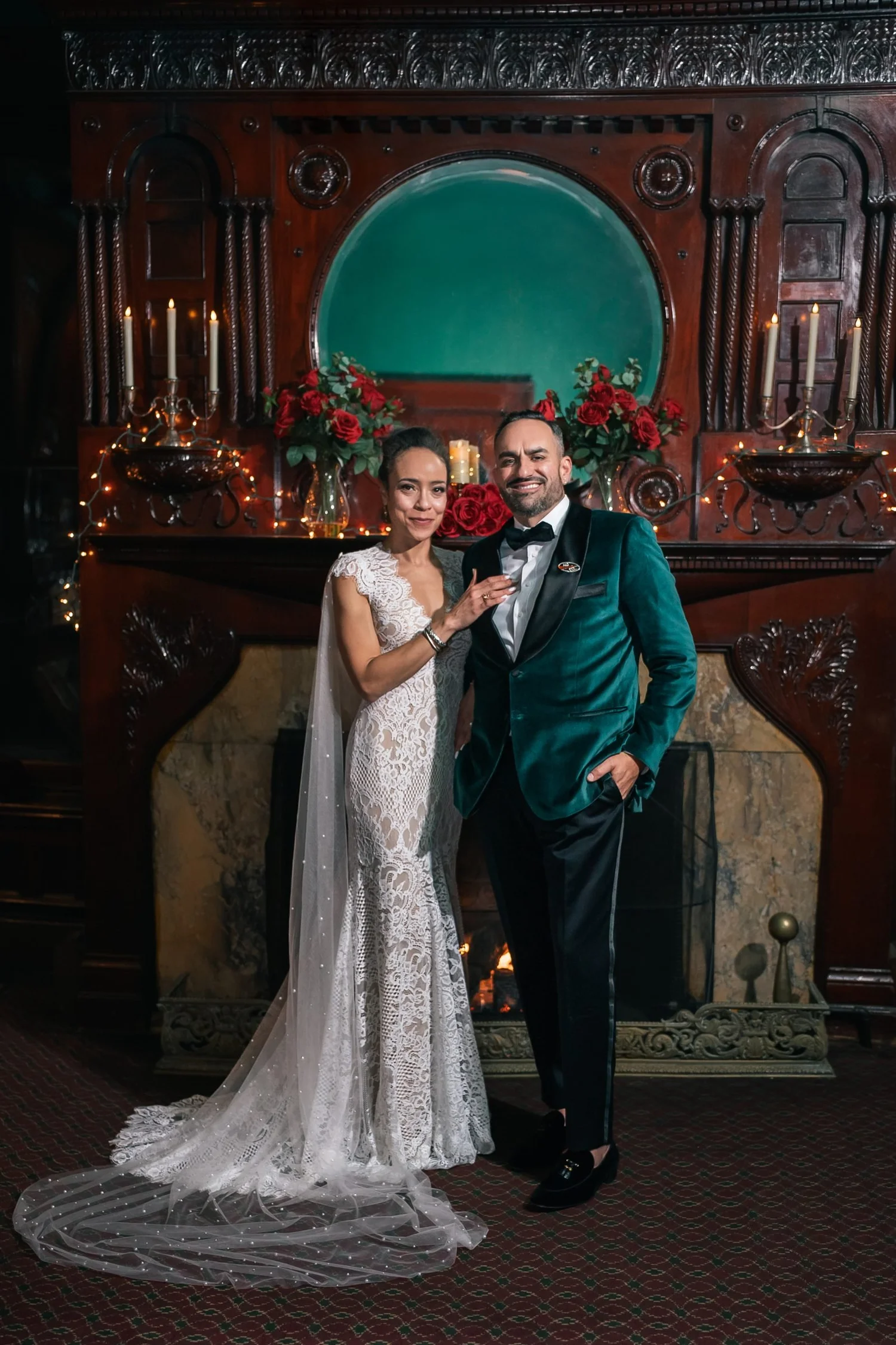Shannon and Leon close together in front of a carved wooden fireplace at Belhurst, with red flowers and candles on the mantel