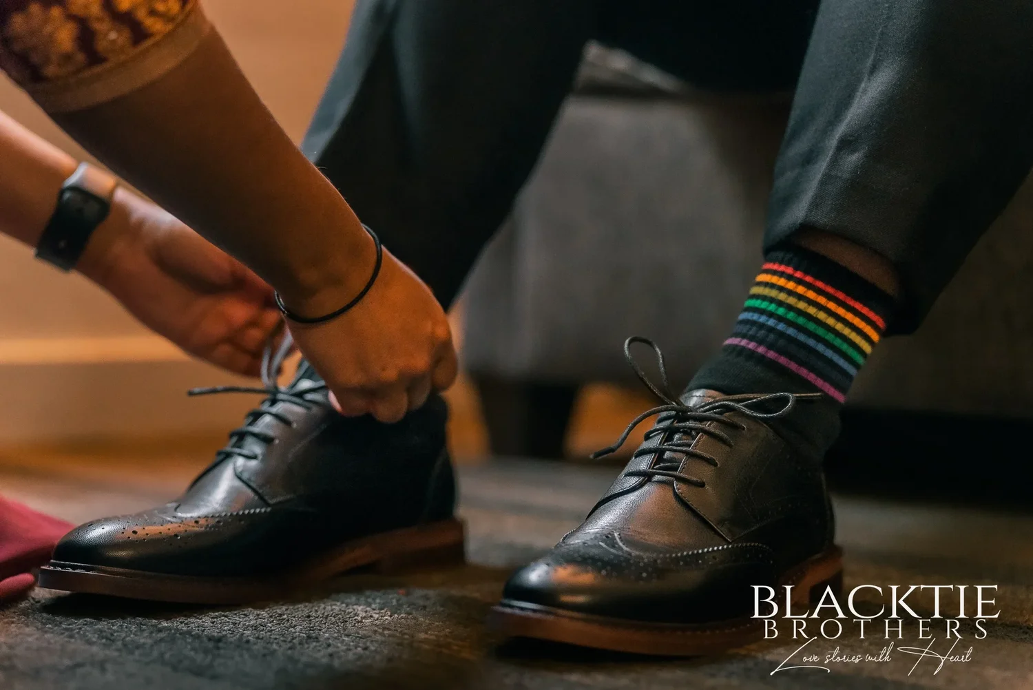 Hands tying the laces of black wingtip shoes over rainbow striped socks